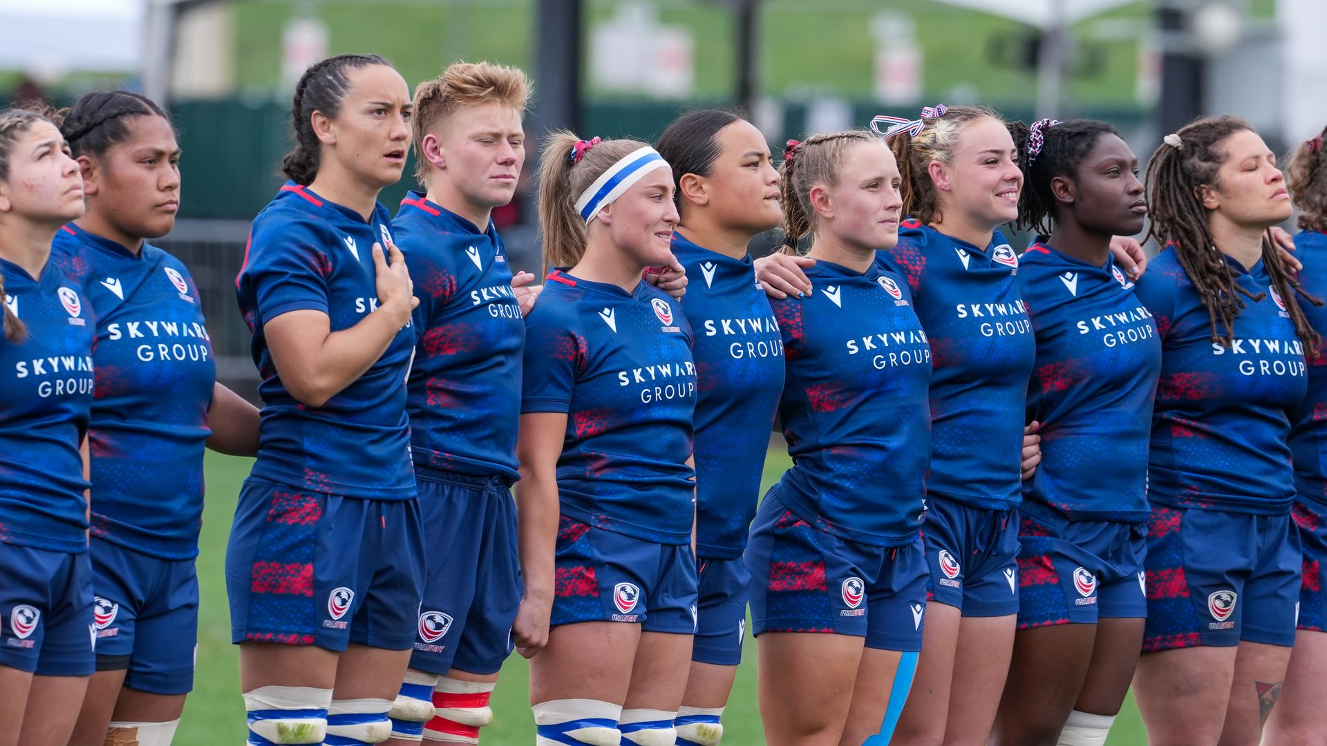 Line of female rugby players in blue uniforms with Skyward Group on their shirts, arms around each other, standing on a grassy field during a team event, focused expressions.