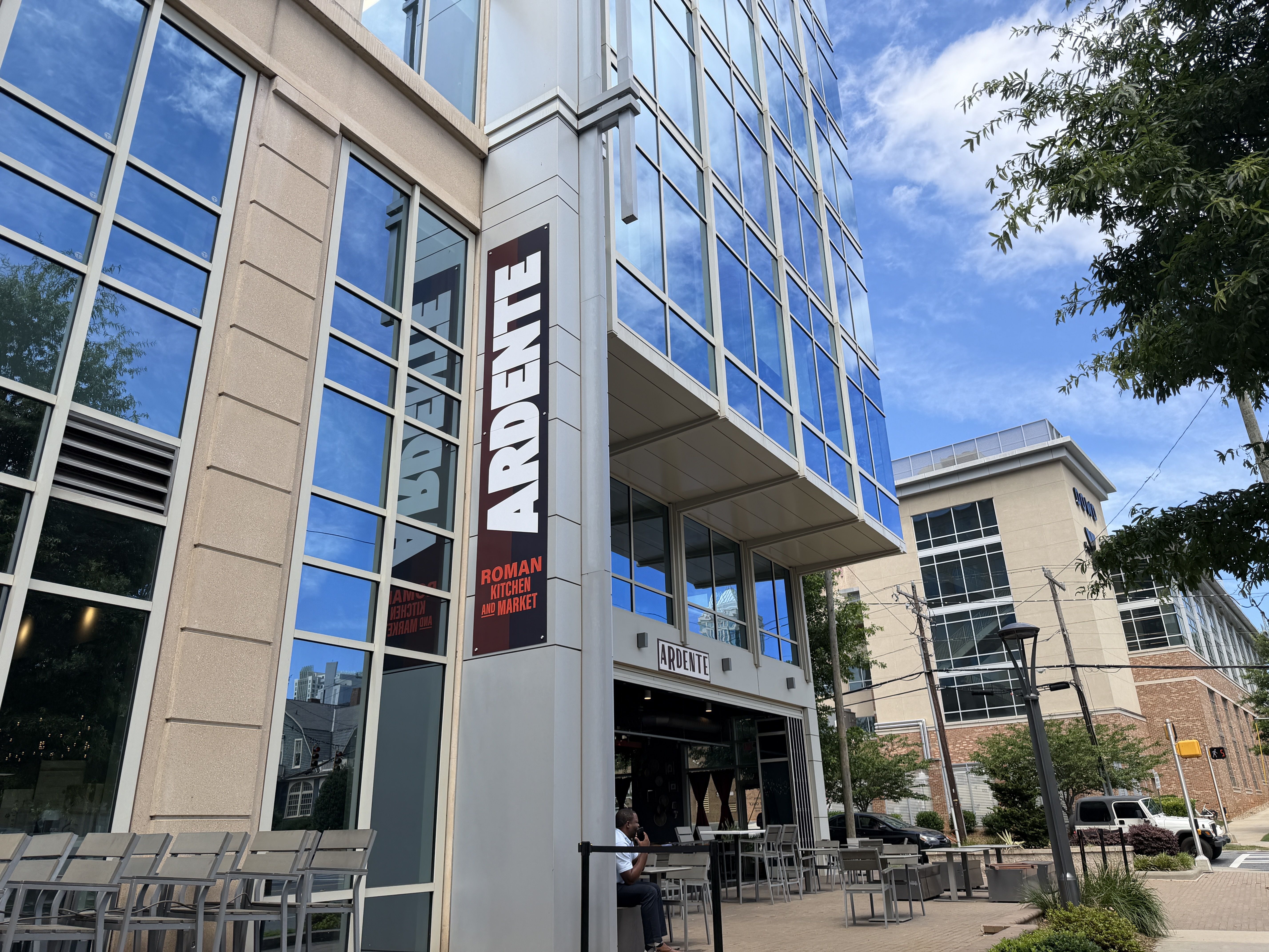 Modern glass-front building reflecting a bright blue sky, with a tall vertical sign displaying "ROMAN" and "ARDENTE" at the entrance; outdoor seating, a tree on the right, and a sunny day.