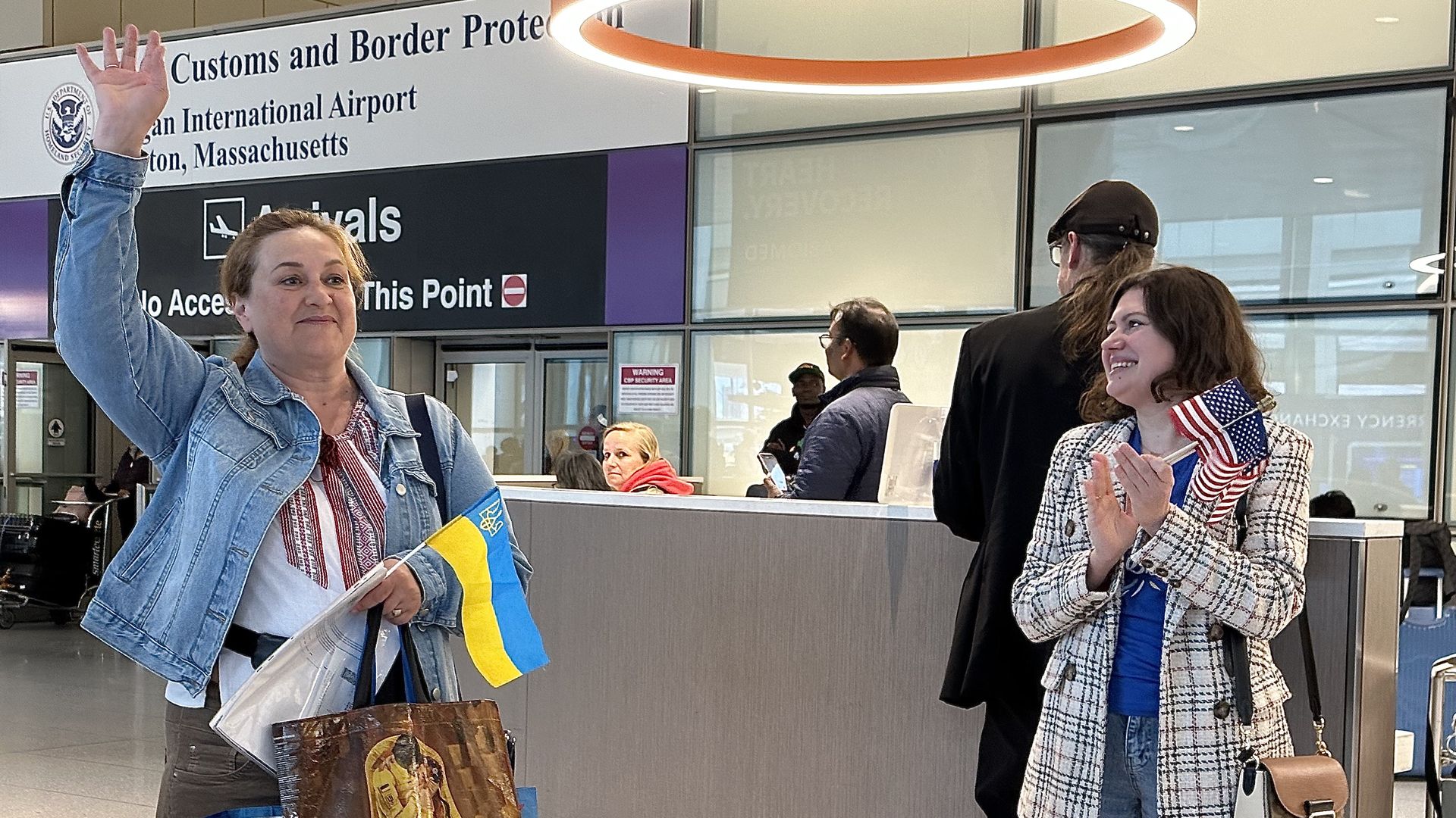 Alina Chernyakova, a Ukrainian refugee, holds her luggage and a small Ukrainian flag as she waves to volunteers at Boston Logan Airport. On the right, is a former refugee who now works for the nonprofit Alight.