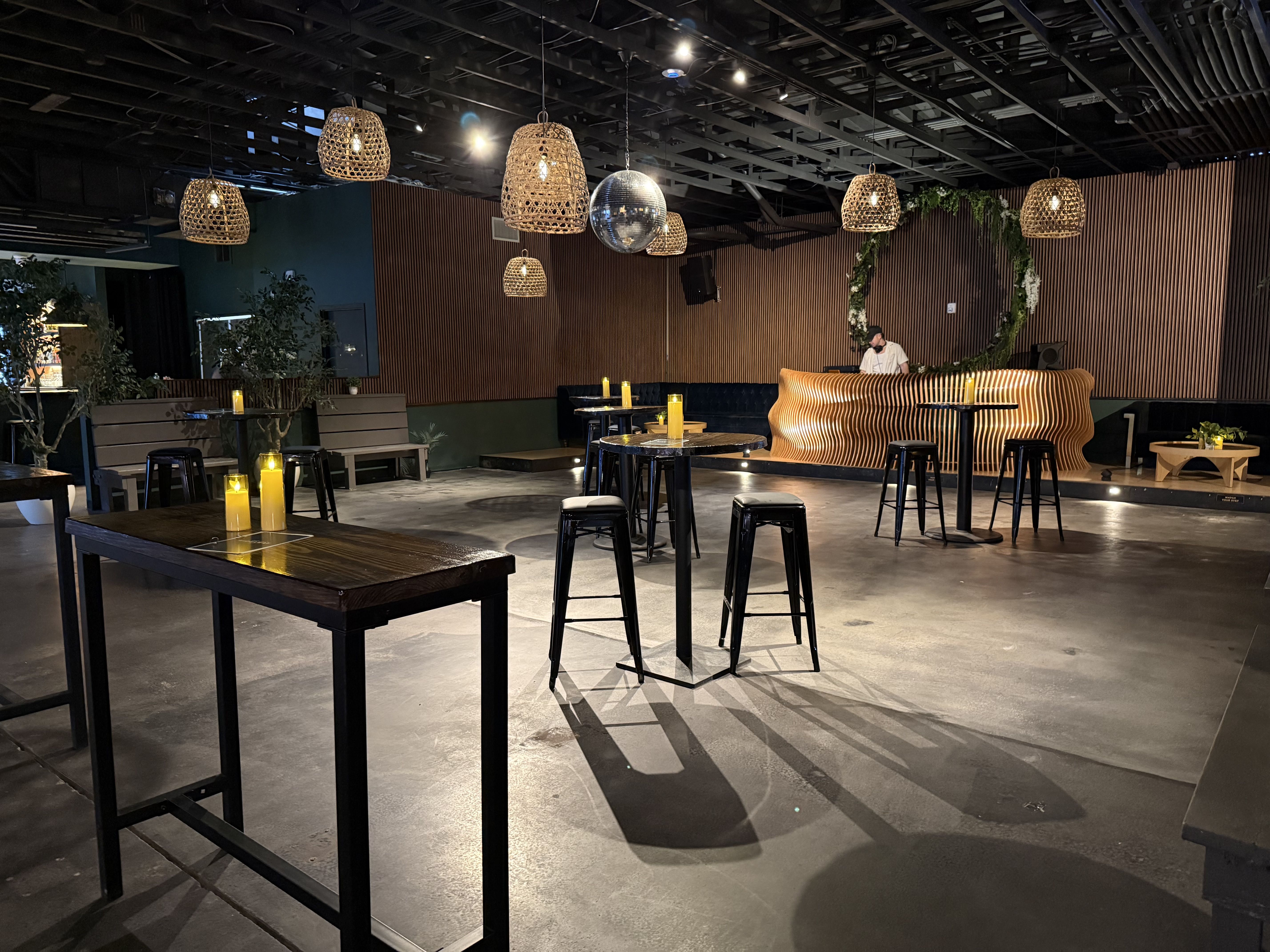 Dim, stylish bar interior with wicker pendant lamps and a central disco ball. Wooden slat walls, a curved orange booth, candlelit tables, black stools, and a person standing near the booth.