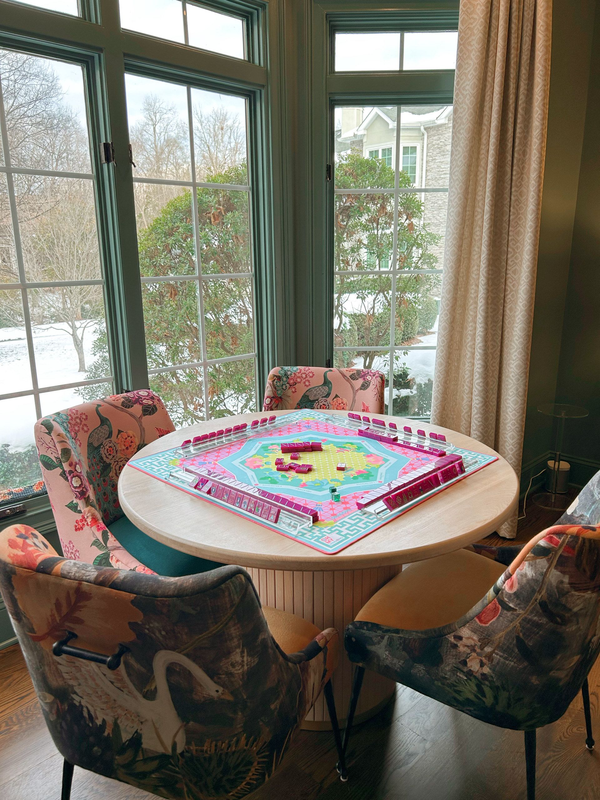 A colorful board game set up on a round wooden table surrounded by four upholstered chairs with floral and bird patterns, next to large windows showing a snowy outdoor scene.