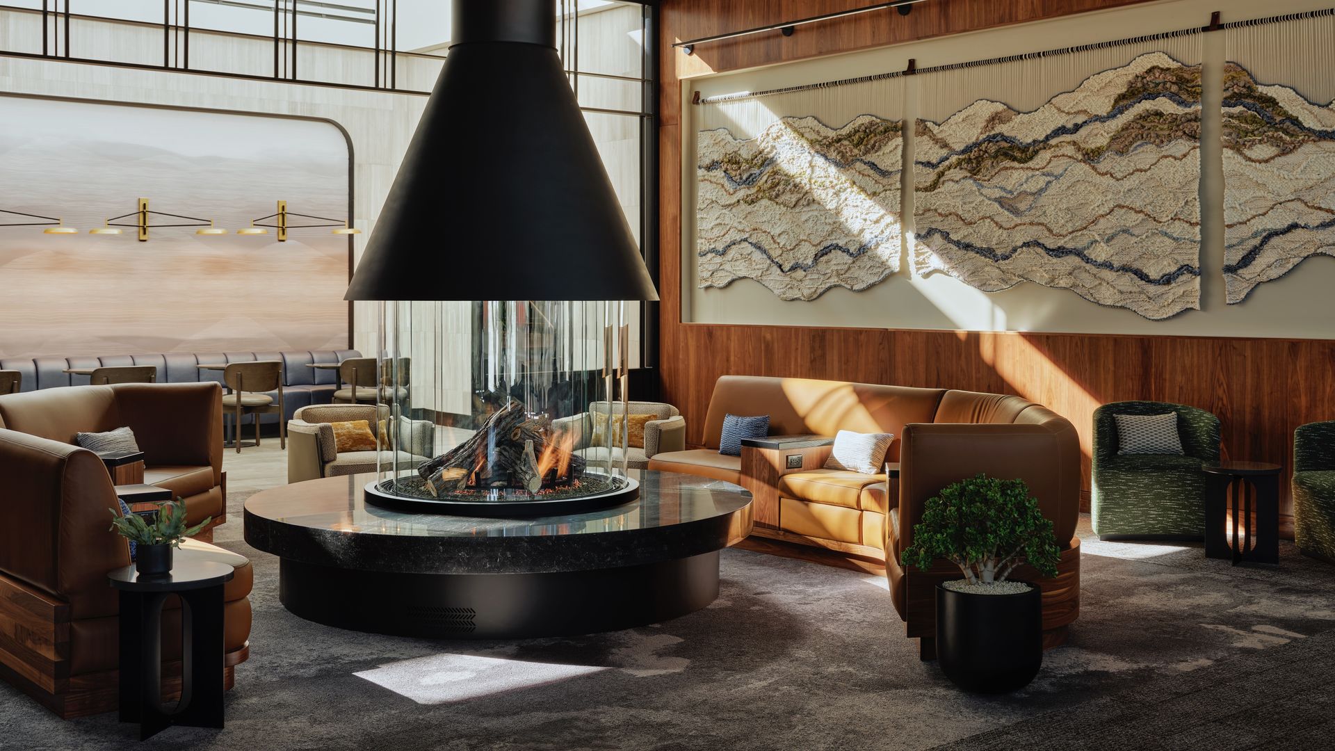 Modern living room with a central black fireplace, brown leather sofas, green armchairs, textured wall art, and large windows letting in natural light.