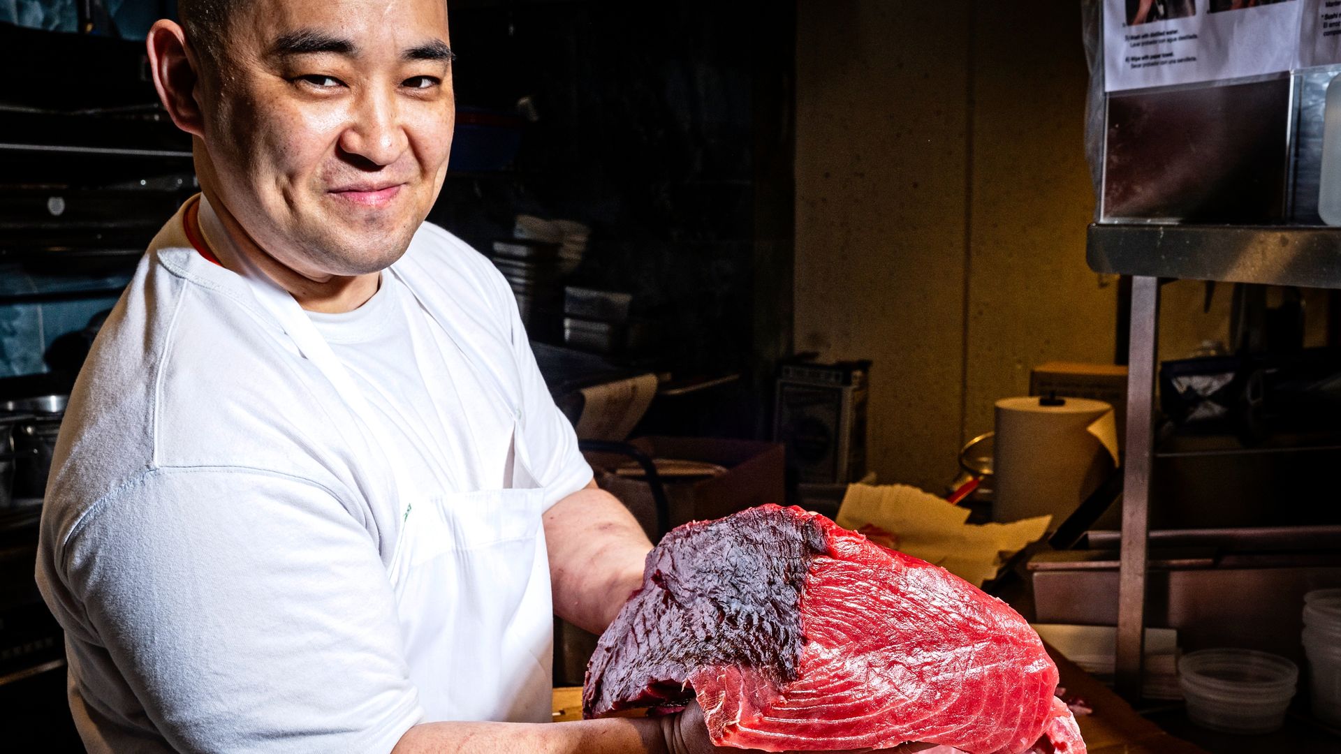 Smiling chef Masaaki "Uchi" Uchino in a white apron holds a large raw tuna cut with dark outer fat, in a kitchen with metal equipment.