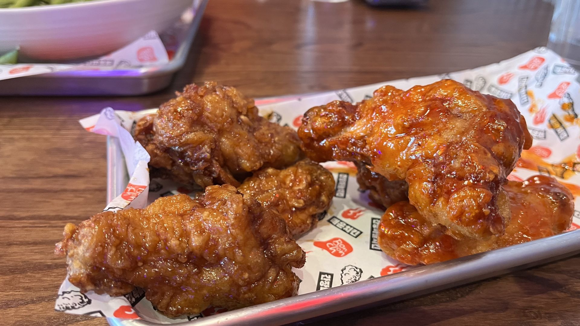 Close-up of glazed fried chicken pieces on a tray lined with branded paper, sticky orange-brown sauce, crispy batter, on a wooden table.