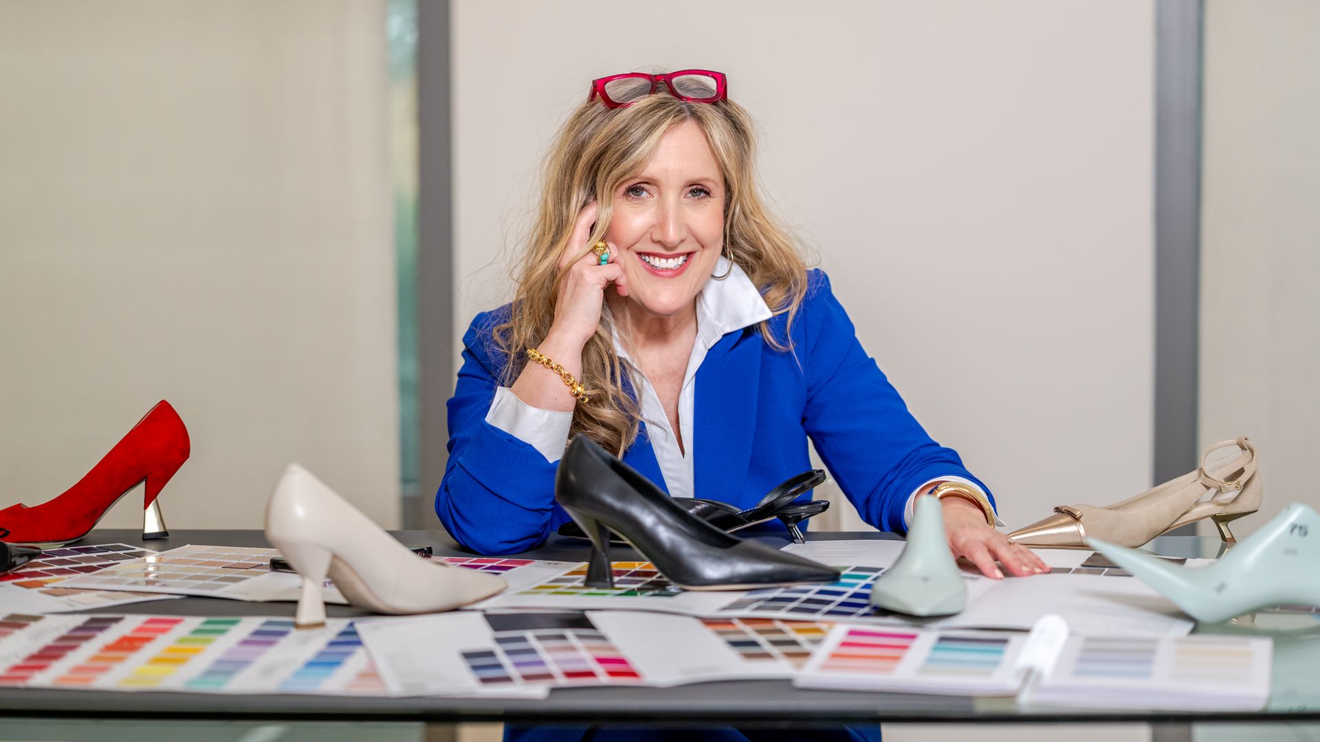 Smiling woman in a blue blazer sits at a cluttered desk with color swatches and many high-heeled shoes, red glasses perched on her head.