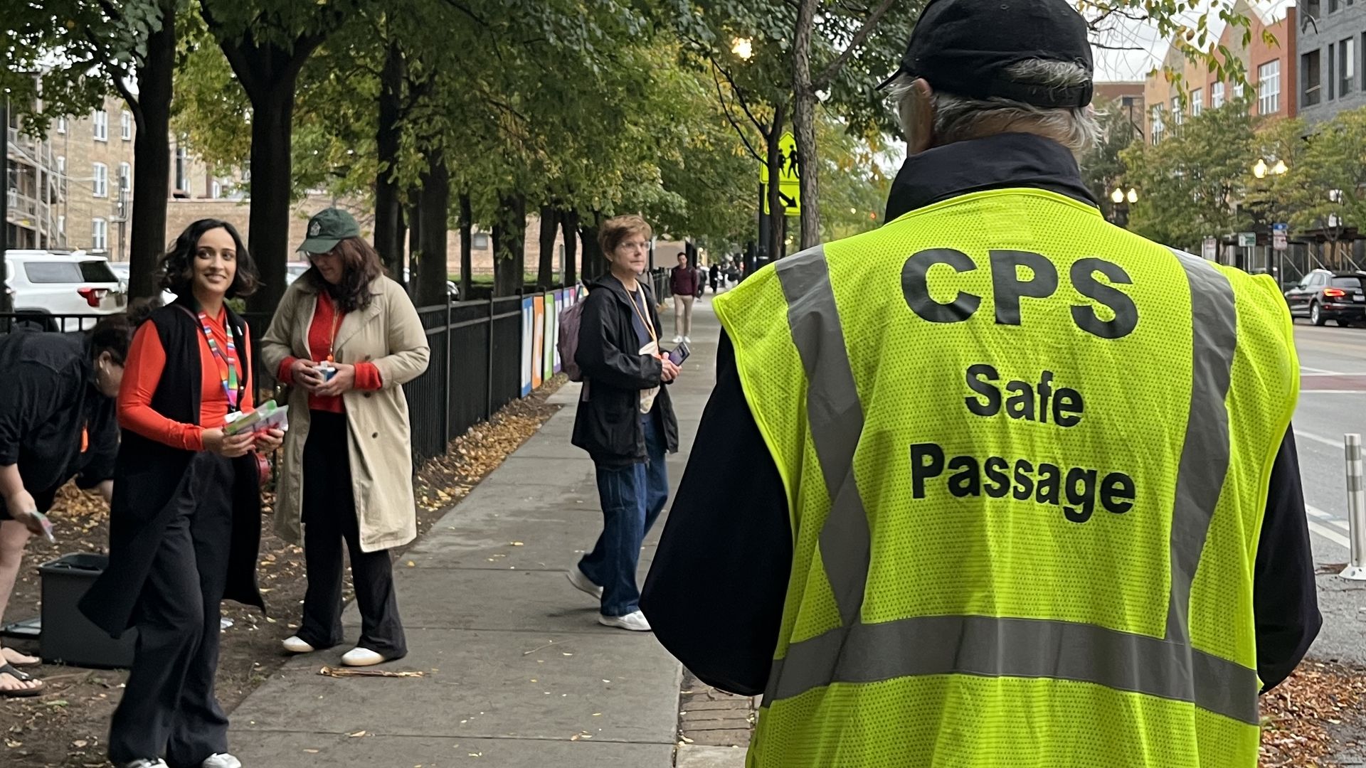 Person wearing a bright yellow safety vest labeled "CPS Safe Passage" stands on a city sidewalk with trees and people walking nearby under overcast sky.
