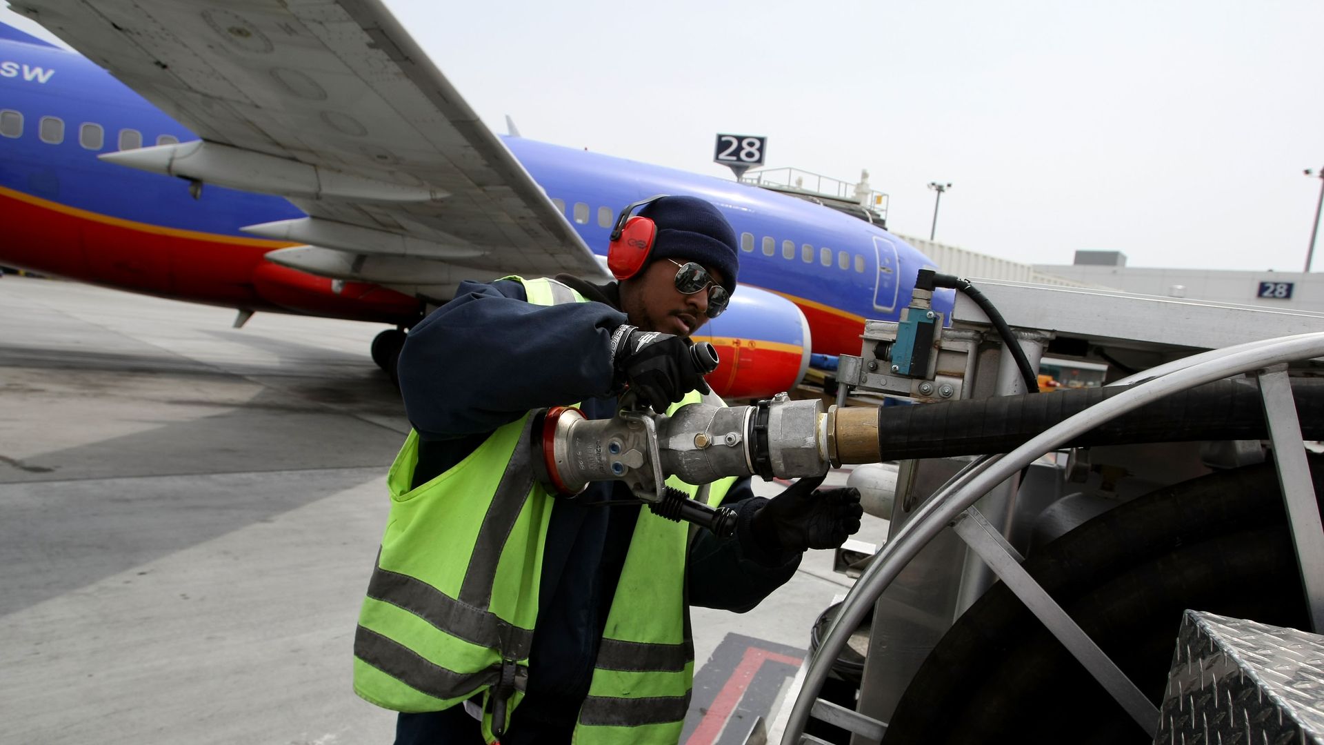 Ground crew member in a neon safety vest, dark jacket, red ear protection and sunglasses operates fueling equipment beside a blue Southwest jet near the wing.