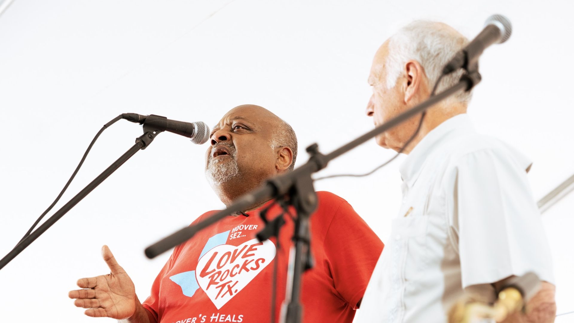 Two older men sing into microphones on a stage; one wears a red T-shirt with a white graphic that says "LOVE ROCKS", the other in a light shirt, against a bright white background.