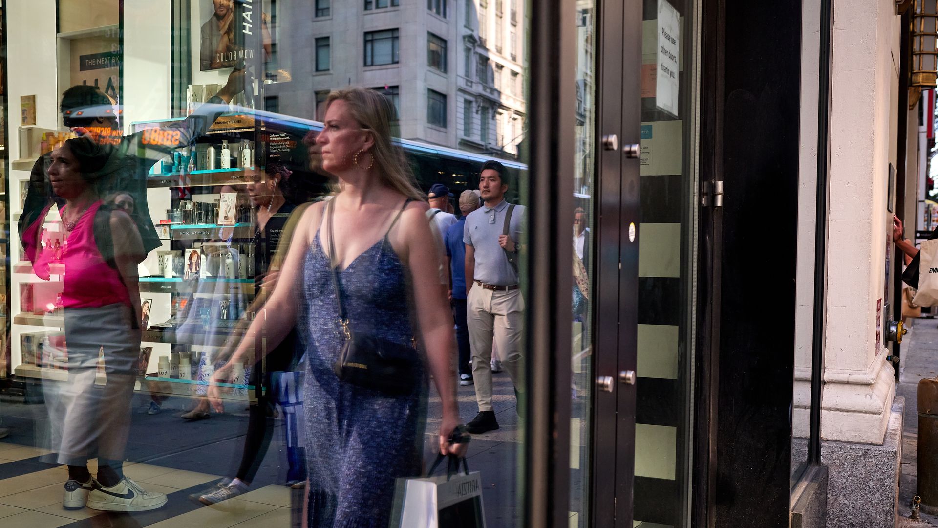 Reflection of pedestrians, including a woman in a blue dress holding a shopping bag, seen through a glass storefront in a busy city with tall buildings and a bus outside.
