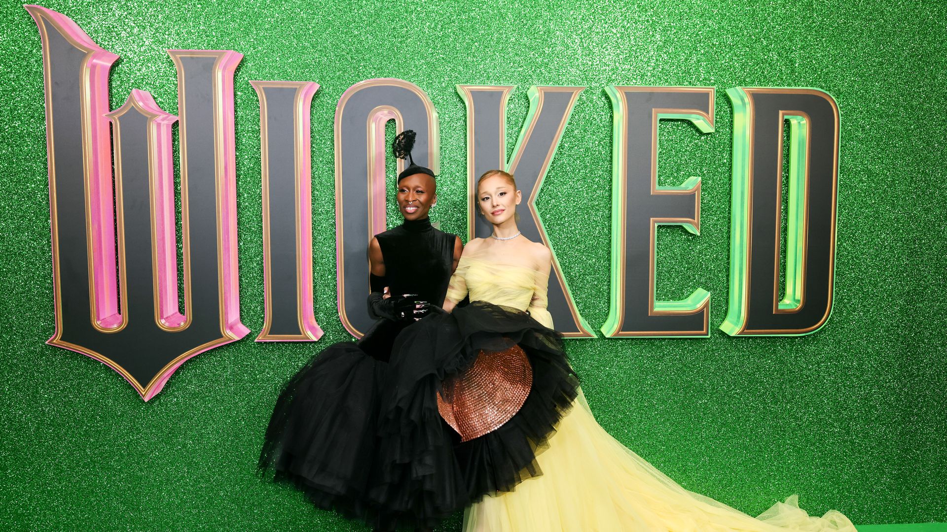 Two women pose in front of a green backdrop that says in black letters, shadowed by pink and green, "Wicked." One woman is wearing a black dress with a tulle skirt. The other is wearing a long pale-yellow gown.