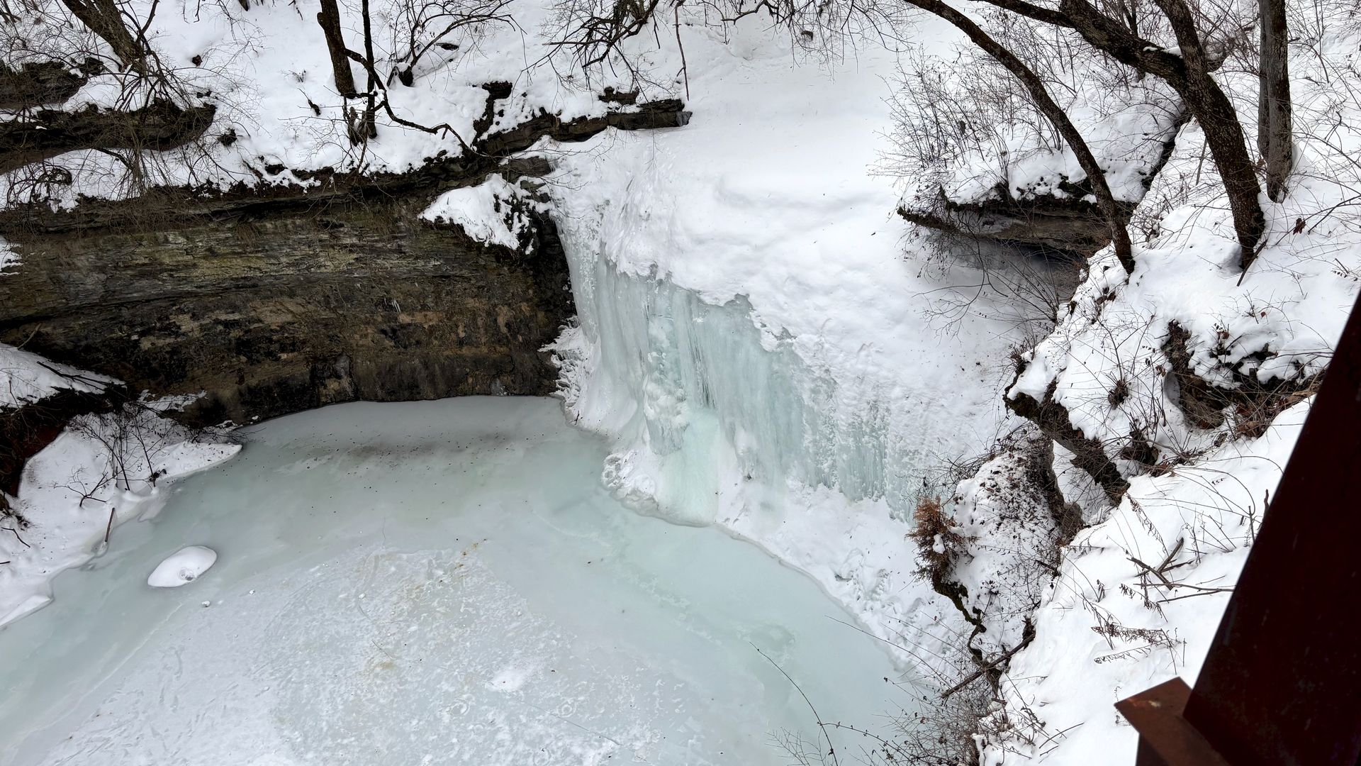 Snow-covered frozen waterfall and river surrounded by bare trees and rocky ledges in a winter landscape.