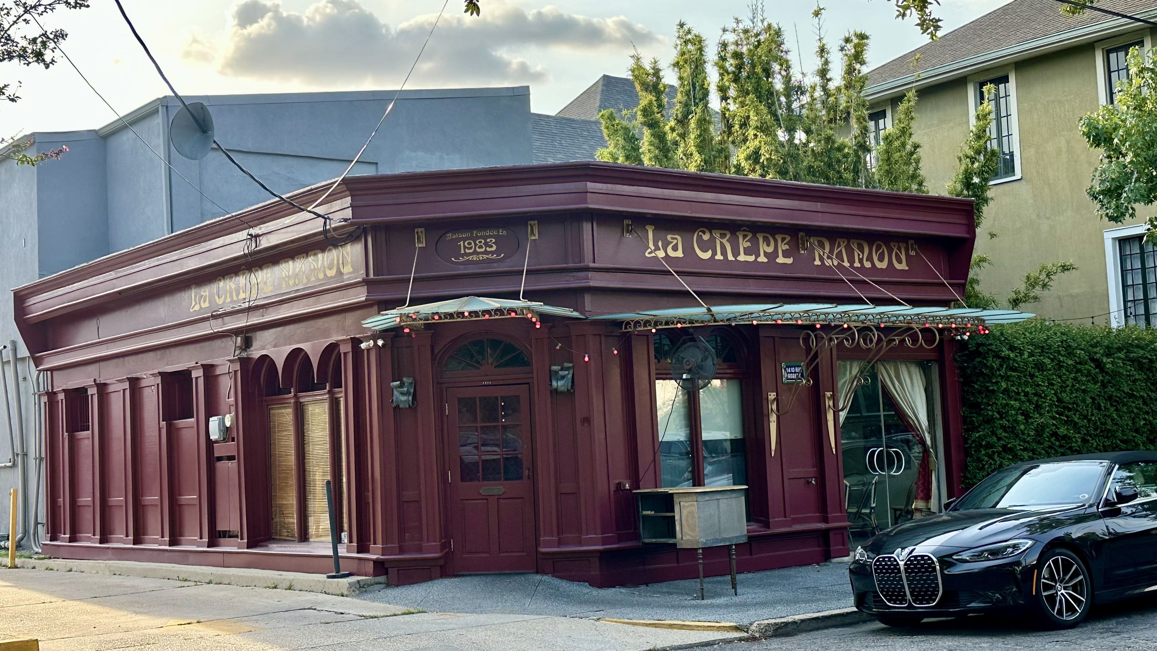 A burgundy storefront cafe named "La Crepe Nanou" with gold lettering, a decorative canopy, string lights, large windows, and a black car parked out front; trees and a beige building behind.