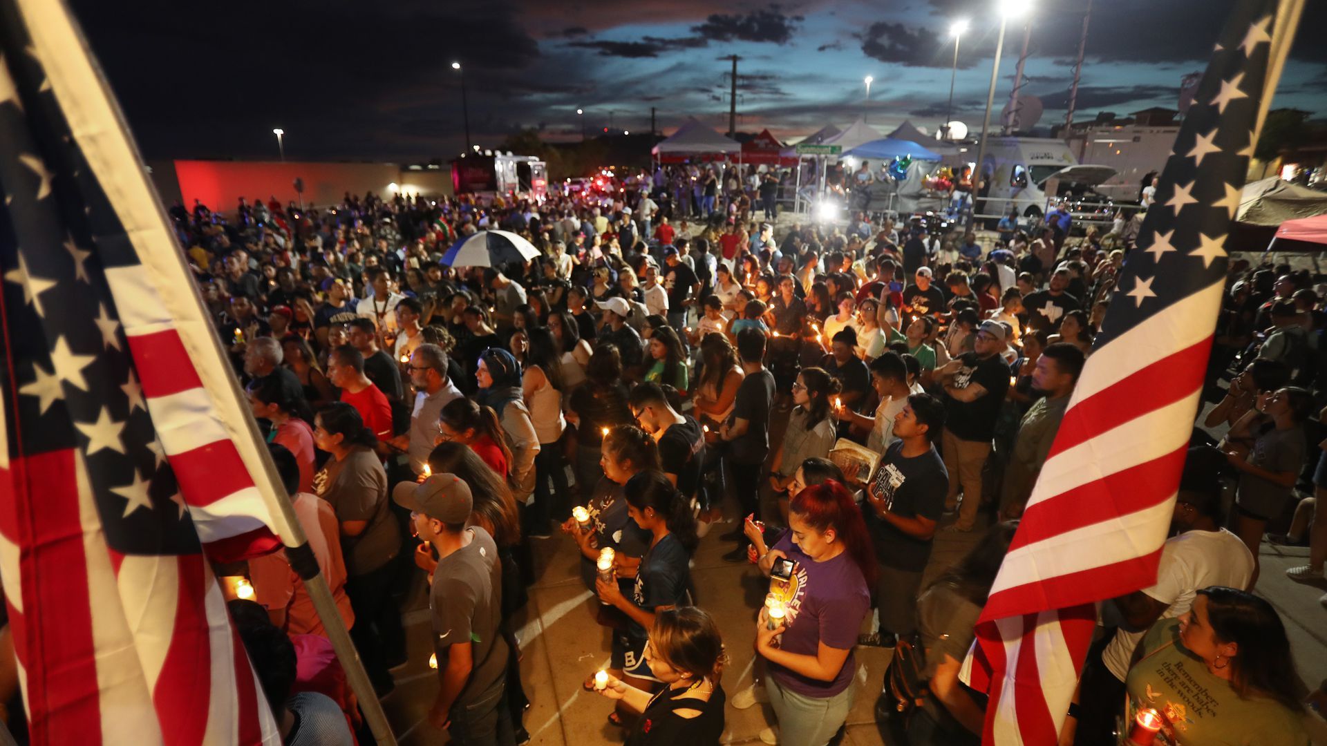 A large crowd of mourners hold candles during a night time vigil in El Paso Texas. one person waves a large American flag. 