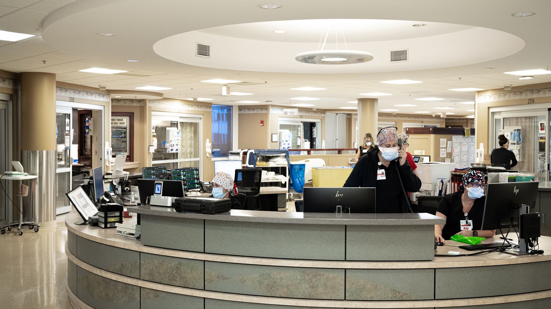 Health care workers sit and walk around a central desk area in a hospital ICU.