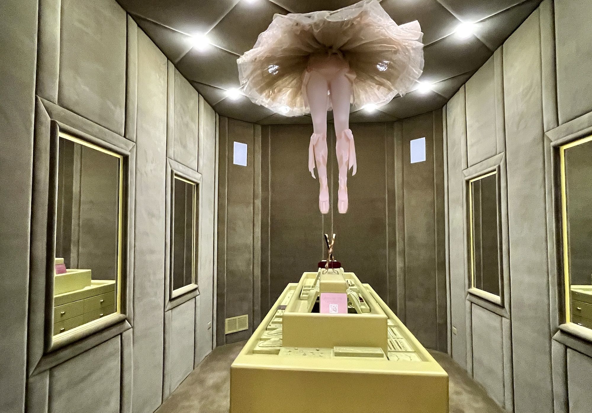 An upside-down figure in a pale pink tutu and tights hangs from the ceiling inside a padded showroom, legs extended downward in pink ballet slippers, under bright overhead lights.