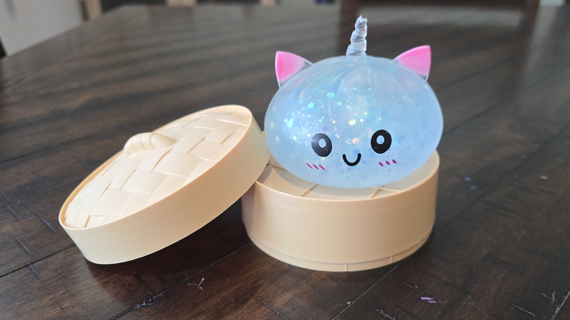 A translucent blue unicorn squishy ball with pink ears and a spiraled horn, glitter specks, black eyes and a smiling mouth, sitting in an open, beige, bamboo-patterned container on a dark wooden table.