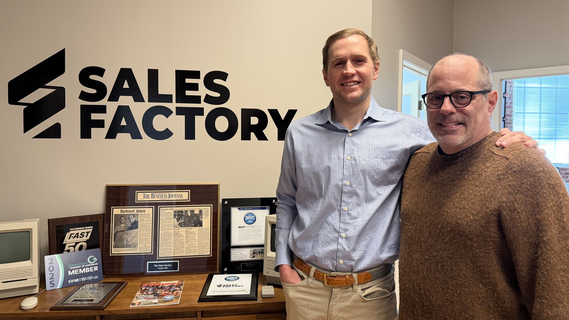 Two smiling men, one in a blue checkered shirt and beige pants, the other in a brown sweater and glasses, standing in an office next to a wall with "Sales Factory" logo and awards on a wooden cabinet.