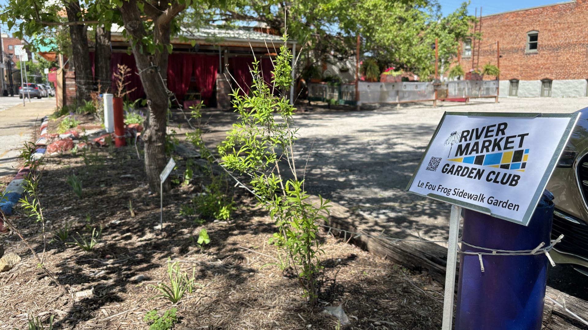 Sidewalk garden display with a sign reading "RIVER MARKET GARDEN CLUB"; foreground shows green shrubs and mulch, while a brick building and a shaded area with red curtains appear in the background.