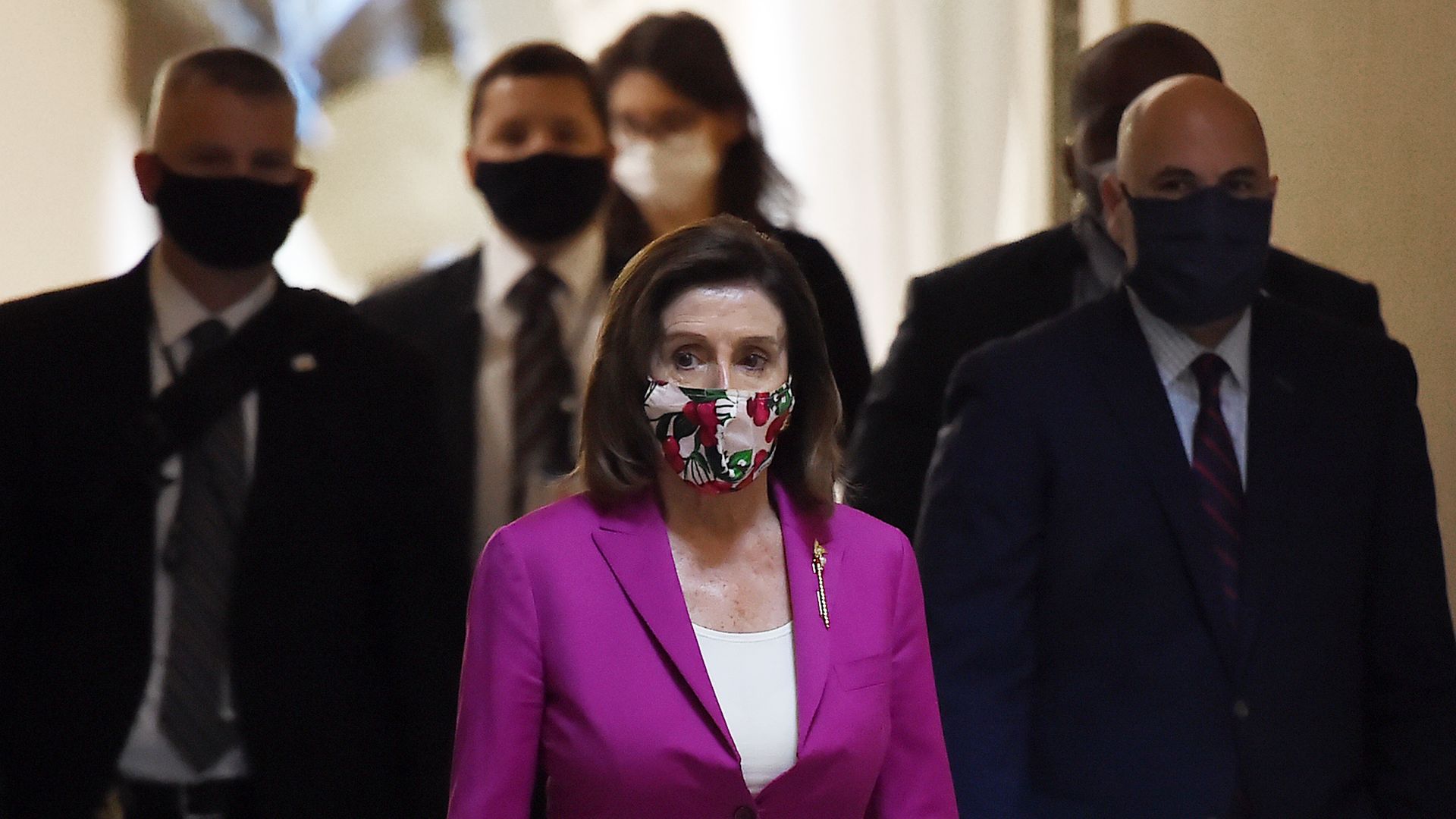 In this image, Nancy Pelosi wears a face scarf while walking ahead of men in suits wearing masks