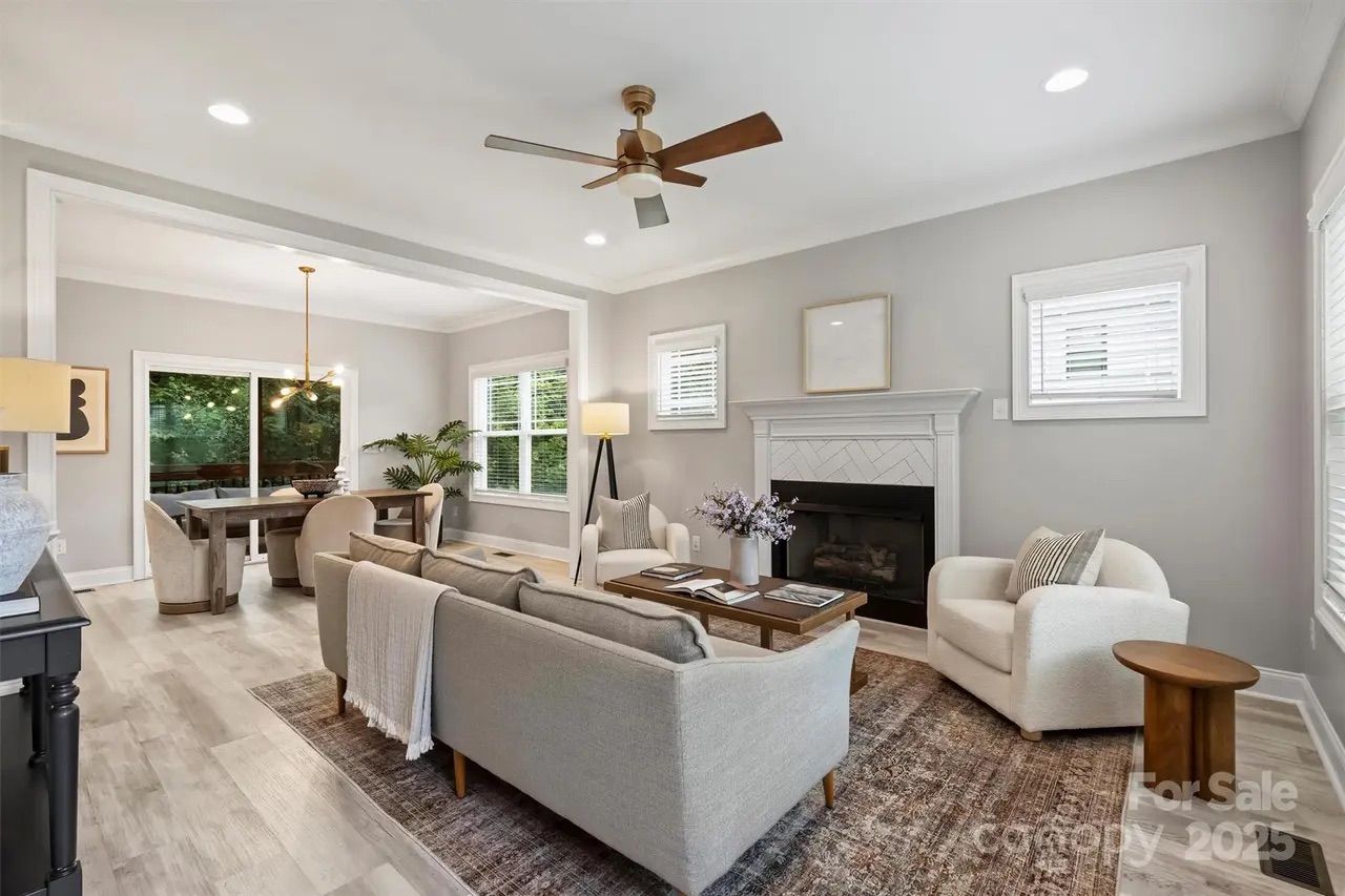 Bright living and dining area with light gray walls, wood floor, beige sofa, two white armchairs, wooden coffee table, fireplace, ceiling fan, and modern chandelier over dining table.