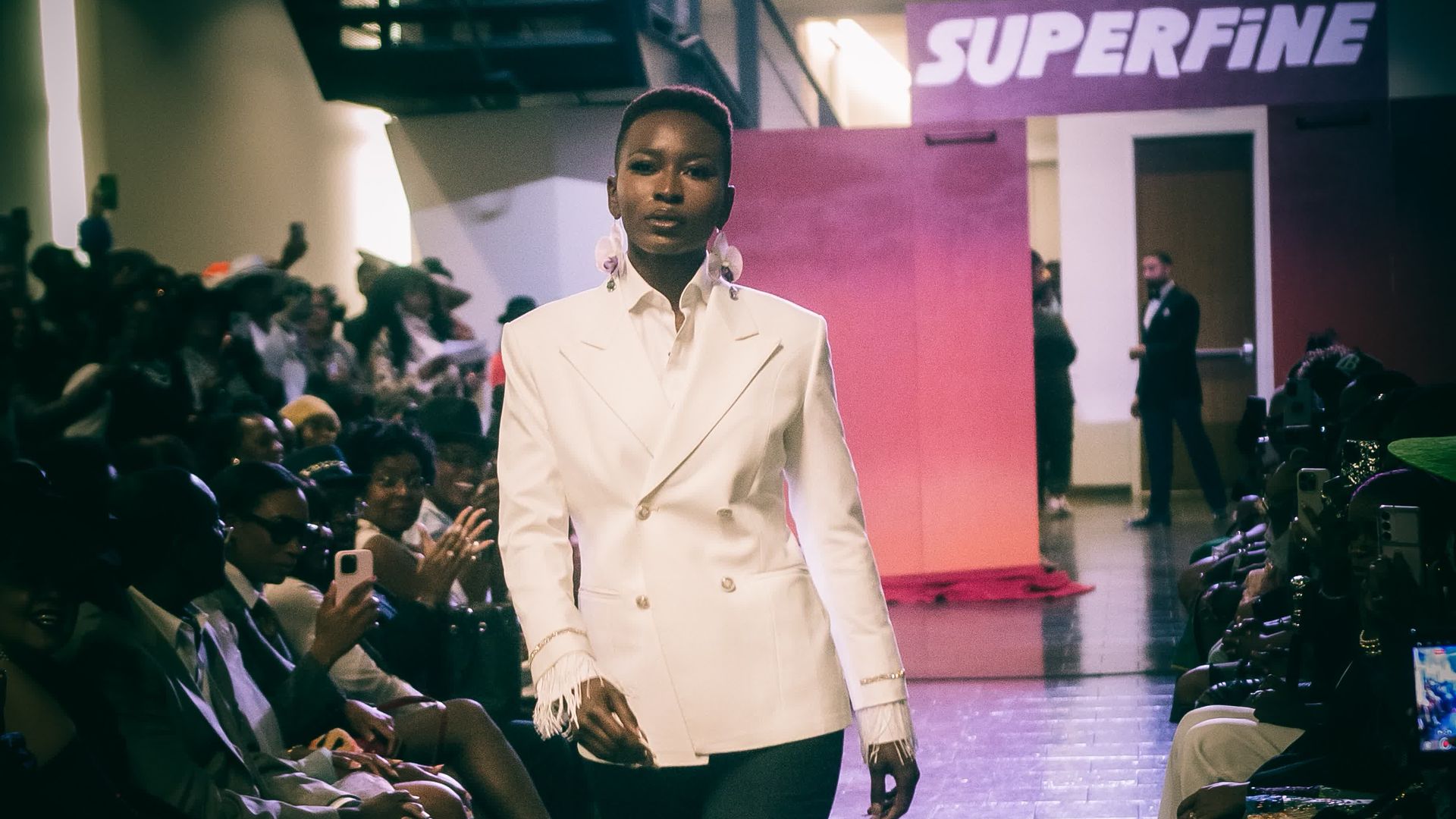 Charlotte's met gala experience. A model walks down a runway wearing a white double-breasted blazer and dark pants at a fashion show with seated audience on both sides and a "SUPERFINE" sign in the background.