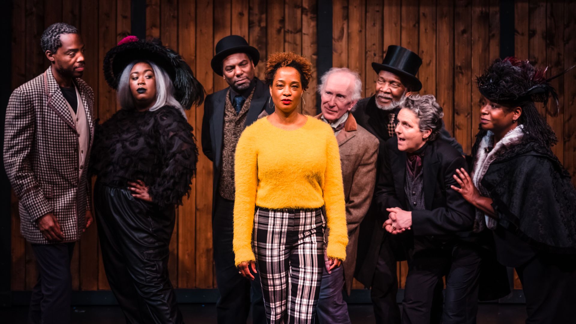 A Black woman in a bright yellow sweater on a stage surrounded by other actors 