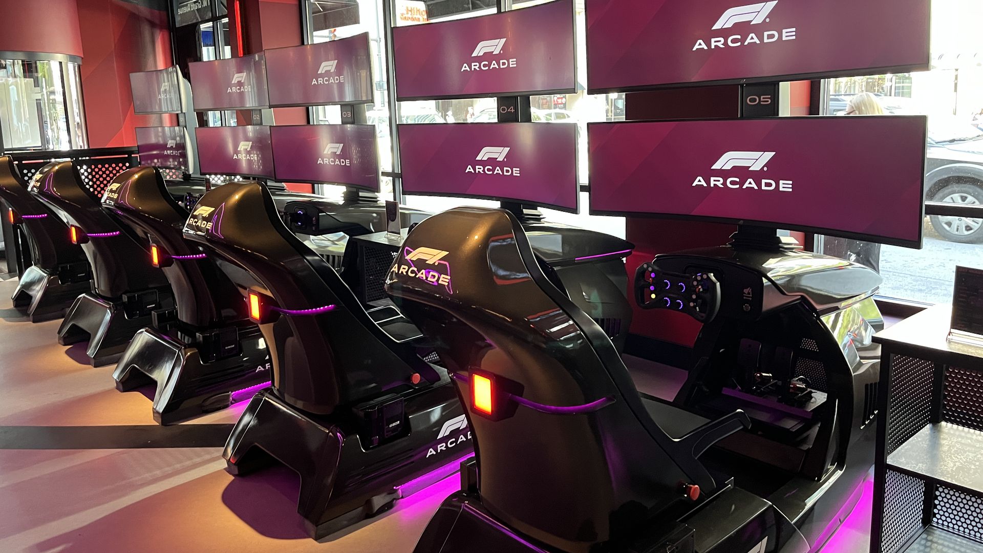 Interior of a neon-lit arcade with a row of black racing simulators, each with a steering wheel and purple underglow. Purple panels read ARCADE beside large windows.