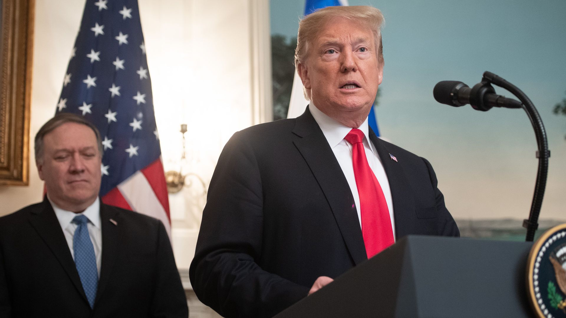 In this image, Trump stands and speaks at a podium as Pompeo stands behind him in front of an American flag.