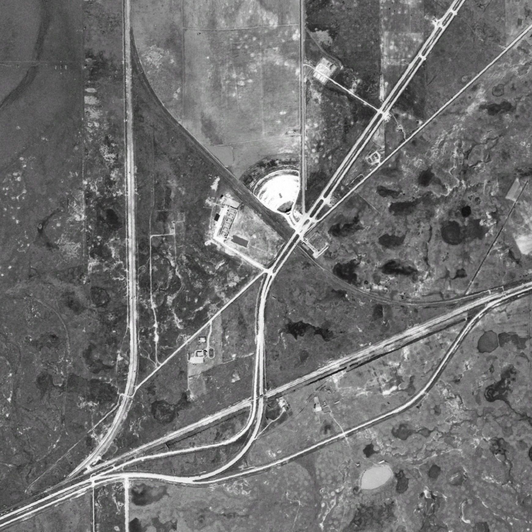 A grayscale aerial view of rural land with a network of intersecting roads; a large circular white pit sits near the center, surrounded by fields, sparse buildings, and scattered vegetation.