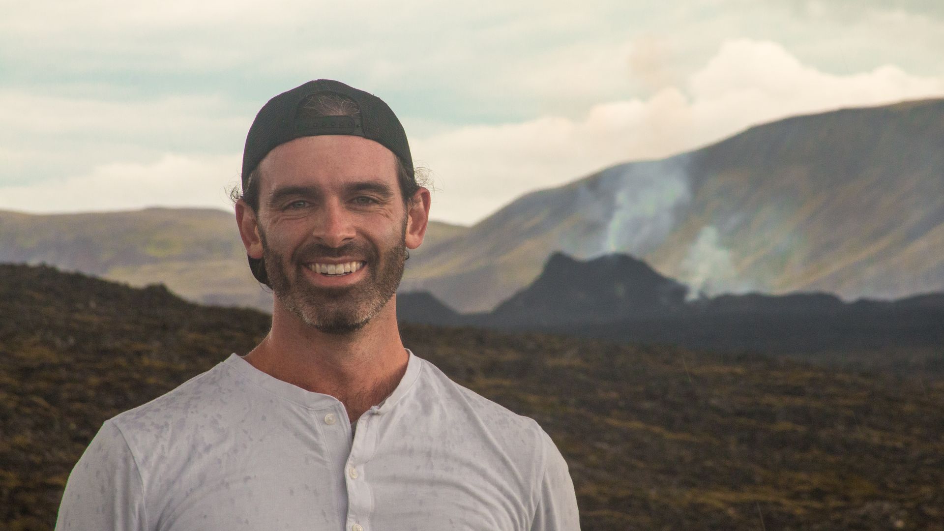 Travis in a white shirt and backwards hat stands outdoors with dark volcanic terrain and smoking volcanic mountain in the background under a cloudy sky.
