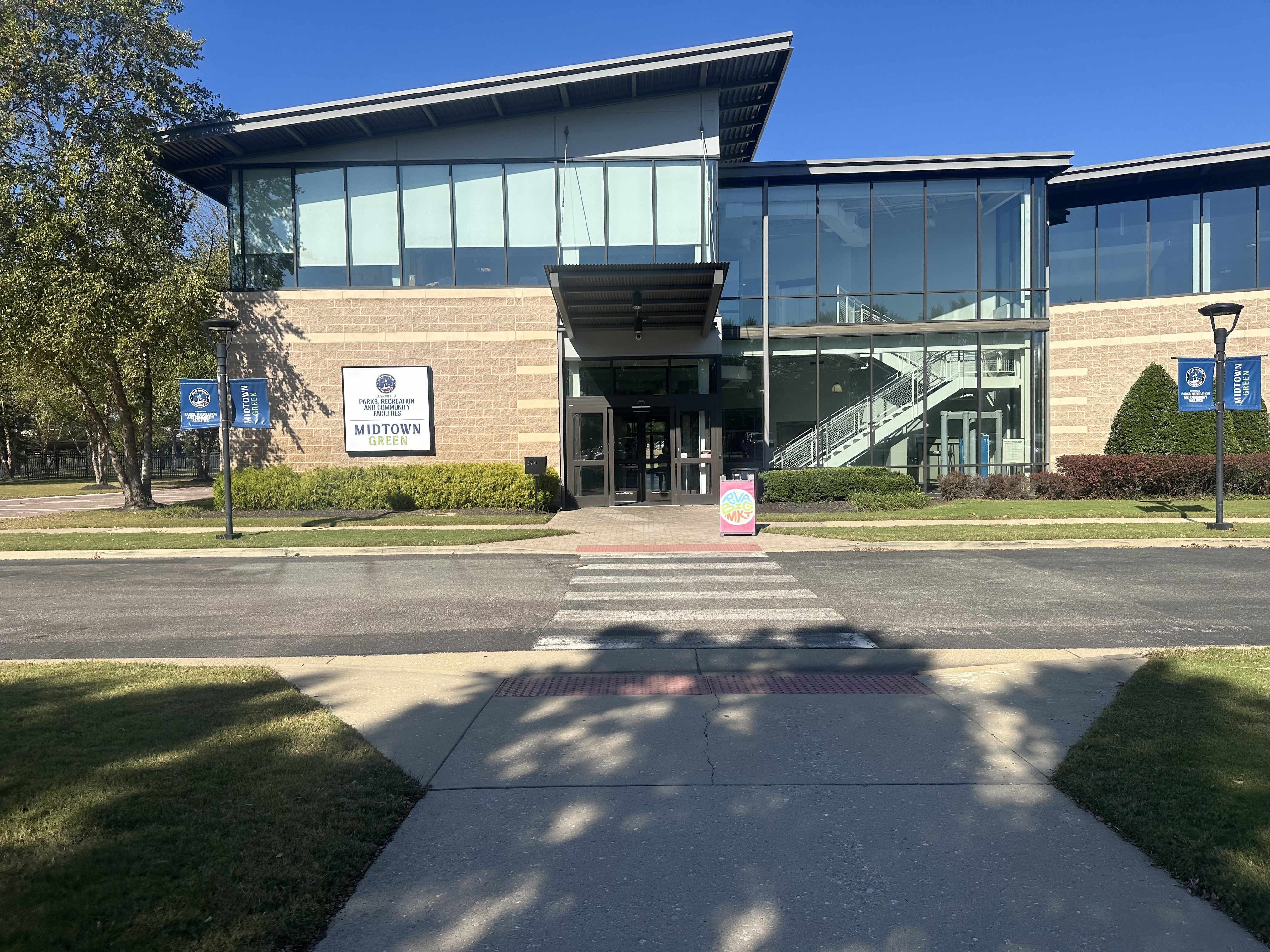 Modern glass-fronted community facility named Midtown Green. Brick lower walls, large blue-tinted windows, metal entry canopy, and visible interior stairs. Blue sky and trees frame the scene.