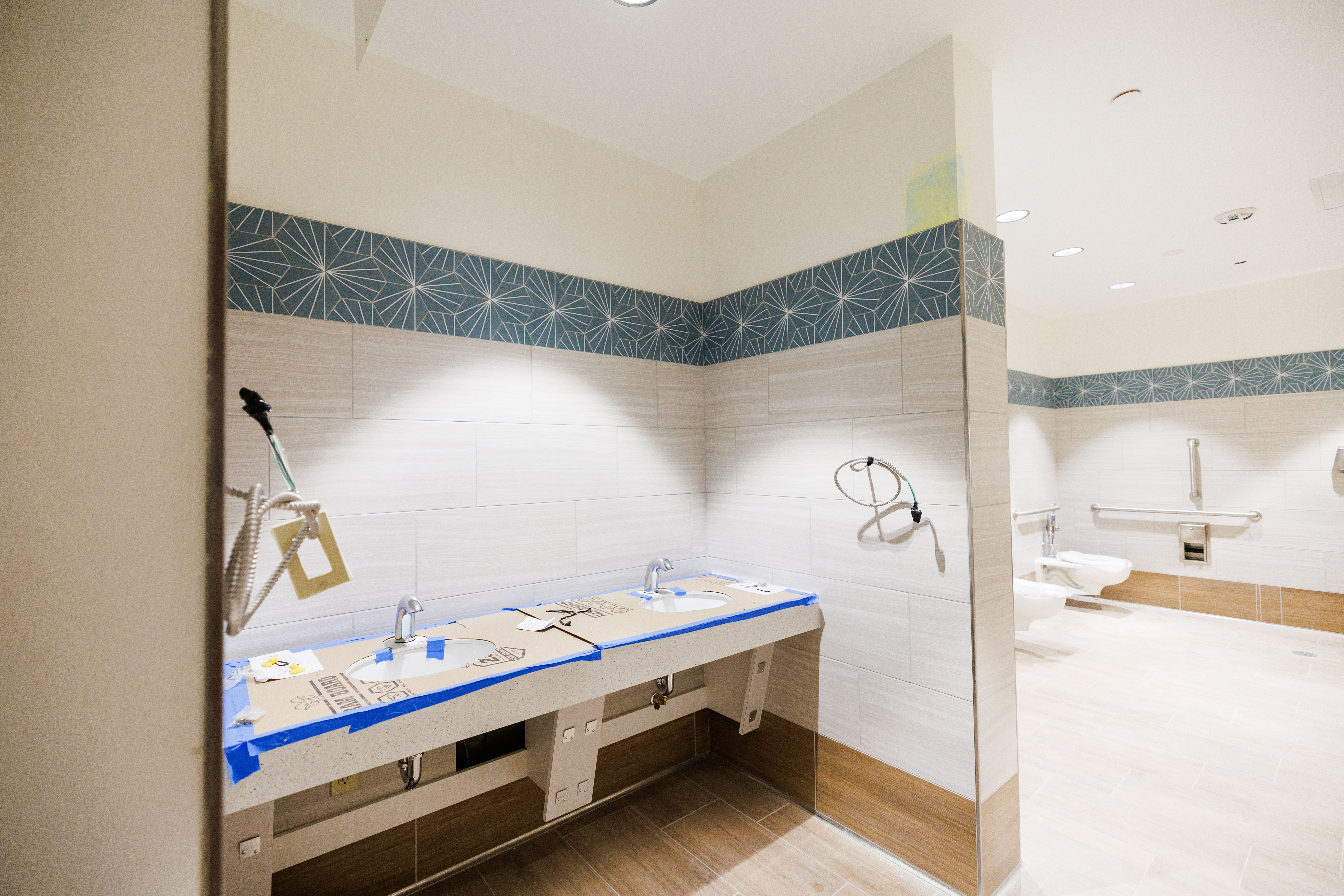 Bathroom sinks with the countertops covered in cardboard, signaling they're not complete yet. In the background are two toilets next to each other. The bathroom is decorated with white, tan and blue tiles.