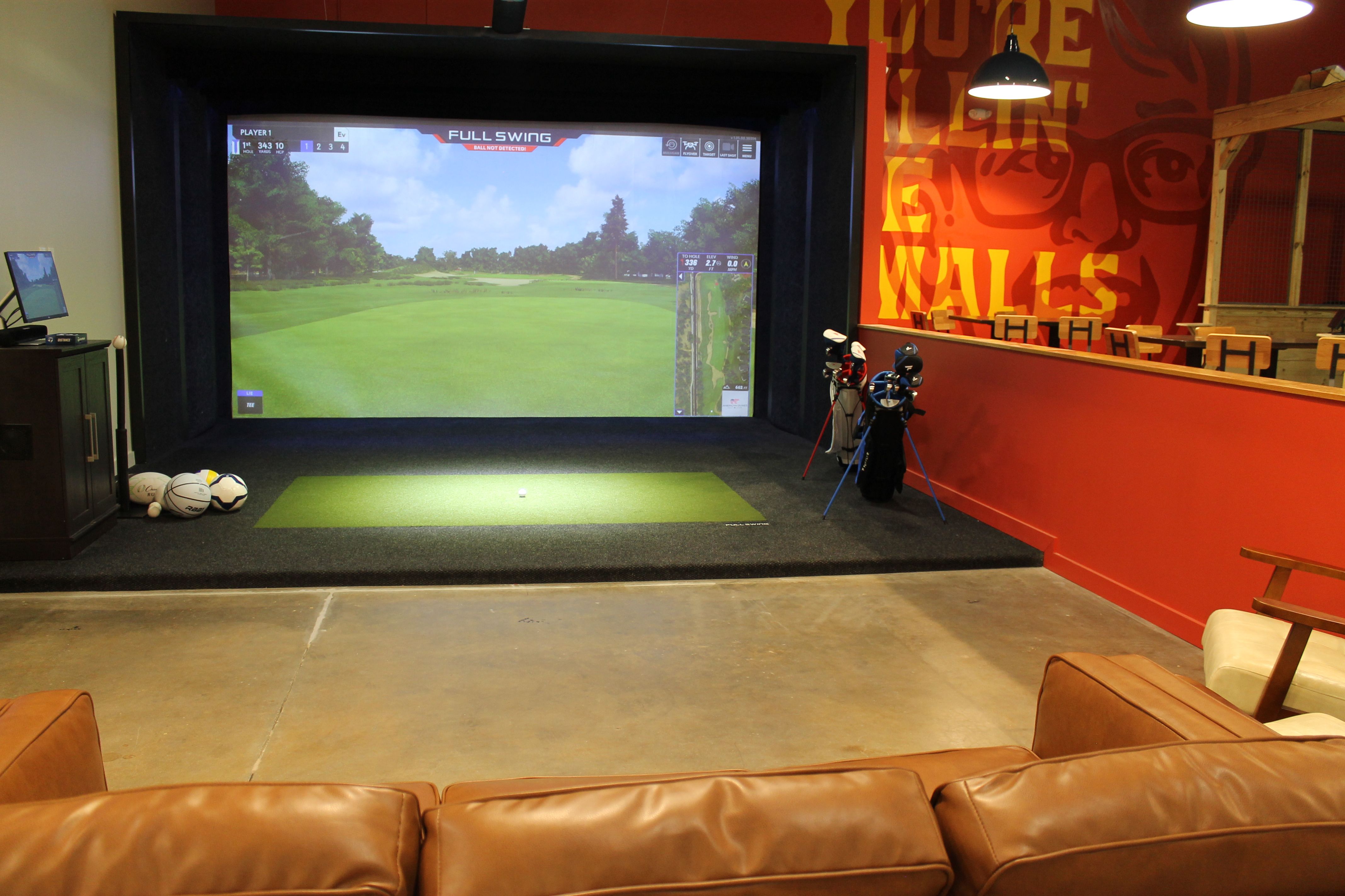 Indoor golf simulator setup with a large screen showing a golf course, golf balls and clubs nearby, surrounded by orange walls with mural text, and brown leather seating in front.