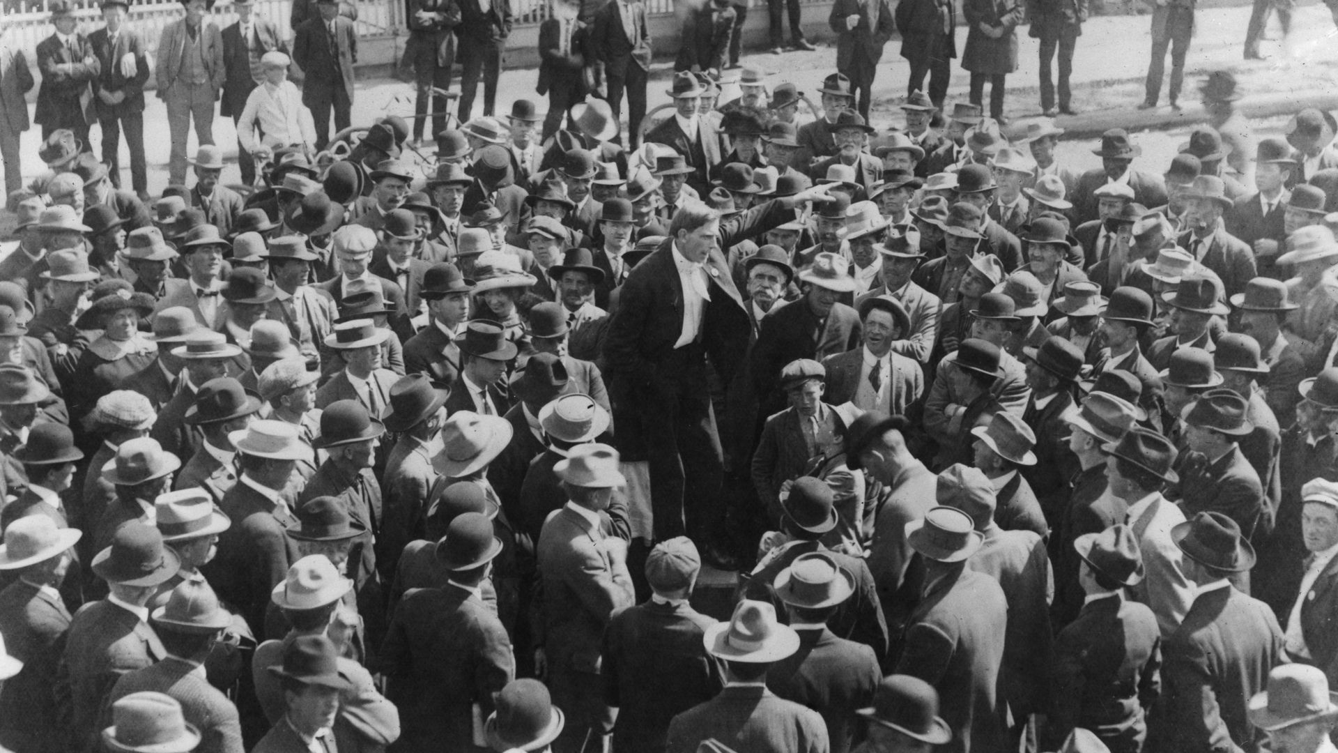 Black-and-white street scene with a dense crowd of men in suits and hats, many bowler hats. Onlookers stand behind a white picket fence; a central man gestures among the crowd.