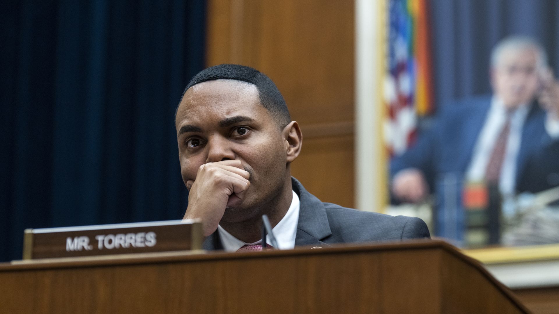 Rep. Ritchie Torres, wearing a gray suit, white shirt and red tie, sitting at a committee dais with his hand over his mouth.