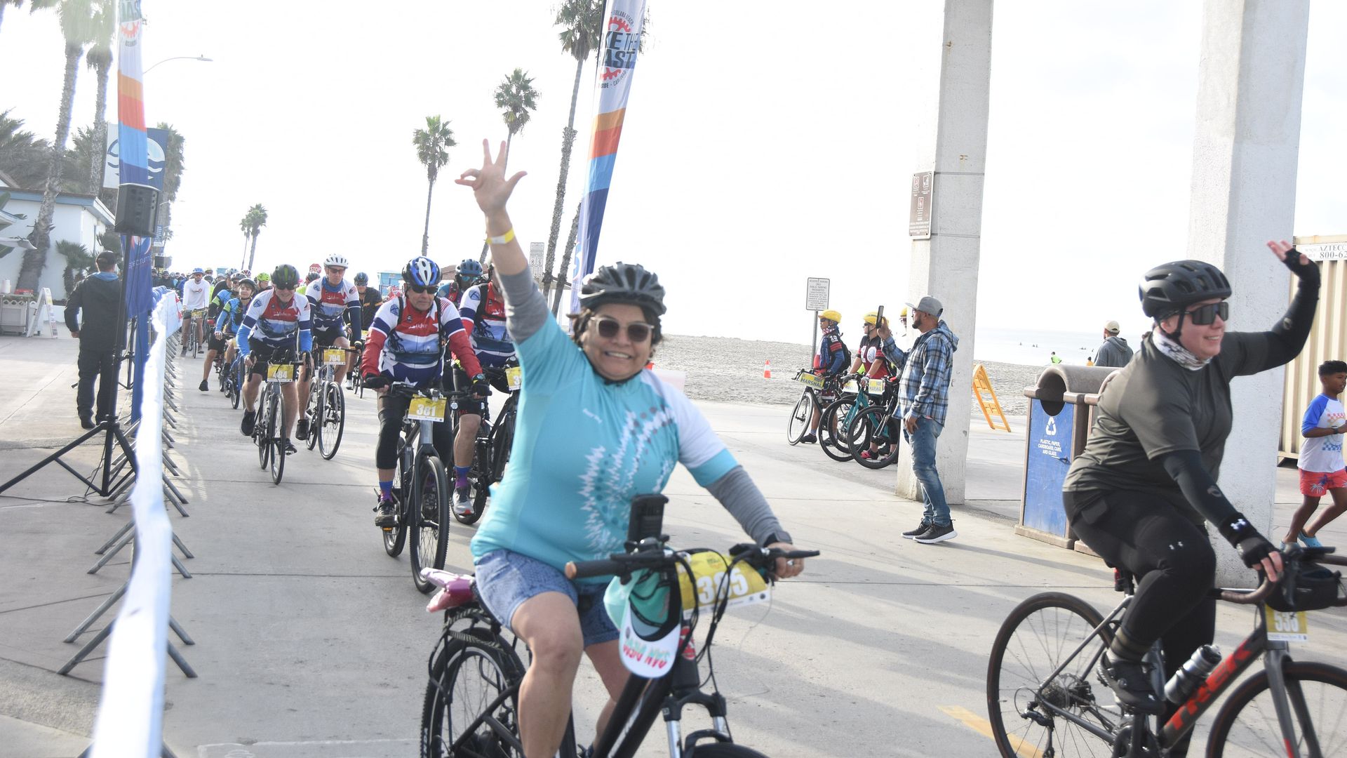 Cyclists in helmets and colorful jerseys riding on a beachside path, two riders in front waving and smiling, palm trees and banners in the background on a sunny day.