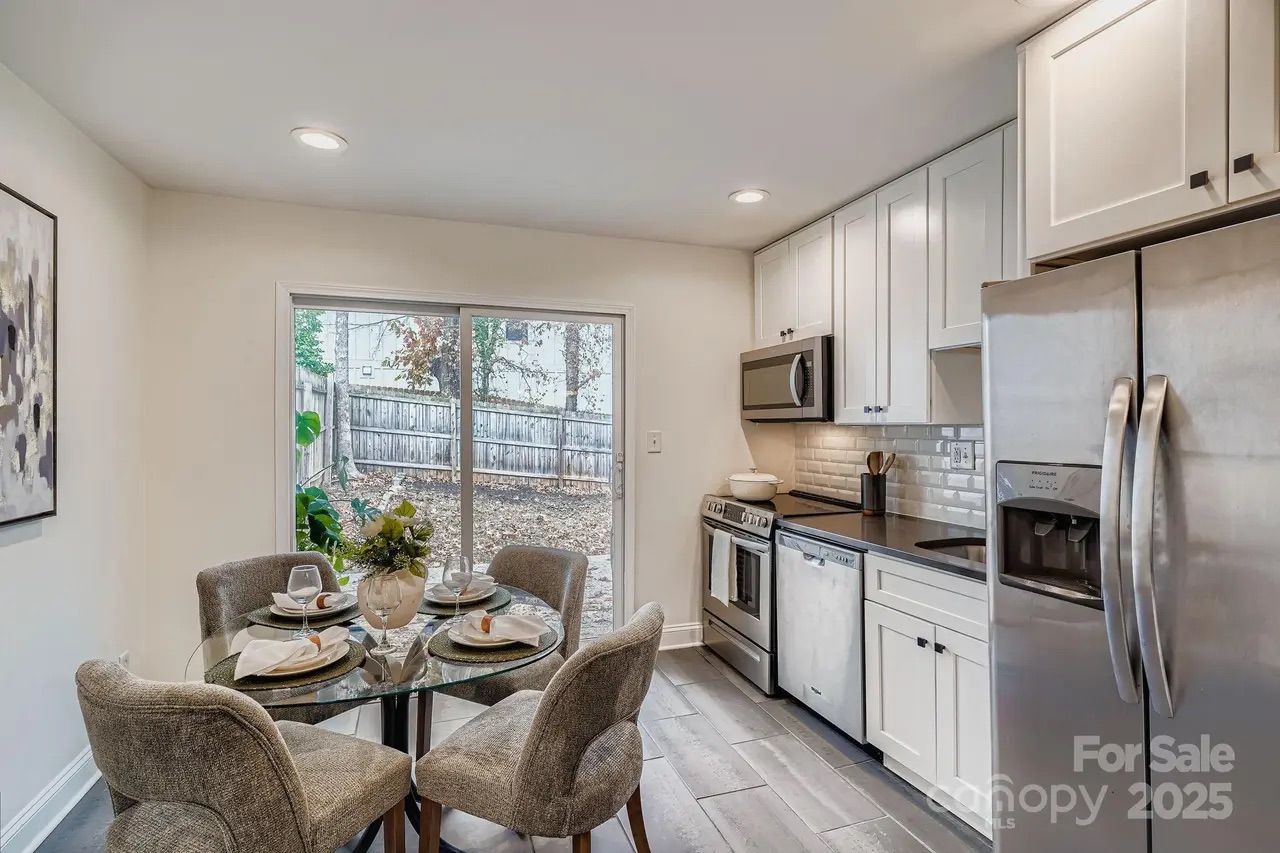 Bright kitchen with white cabinets, stainless steel appliances, glass dining table set for four with gray cushioned chairs, and sliding glass door leading to fenced backyard.