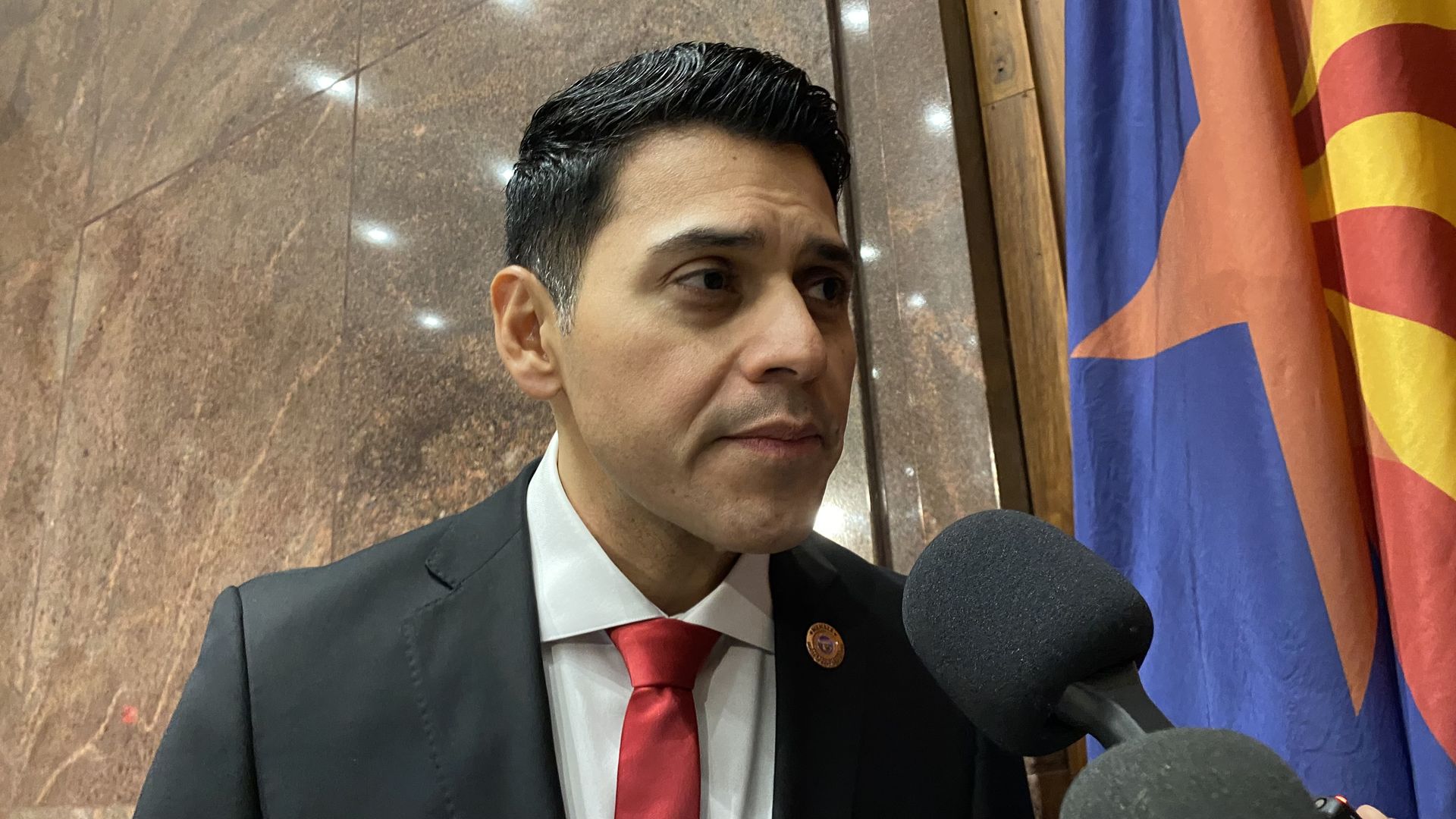 Steve Montenegro speaks into news reporters' microphones with an Arizona flag behind him.