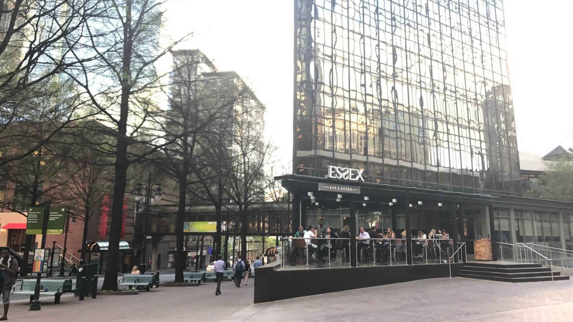 Outdoor dining area of Essex Bar & Bistro with people seated, surrounded by tall trees and modern glass buildings reflecting the evening light in an urban plaza.