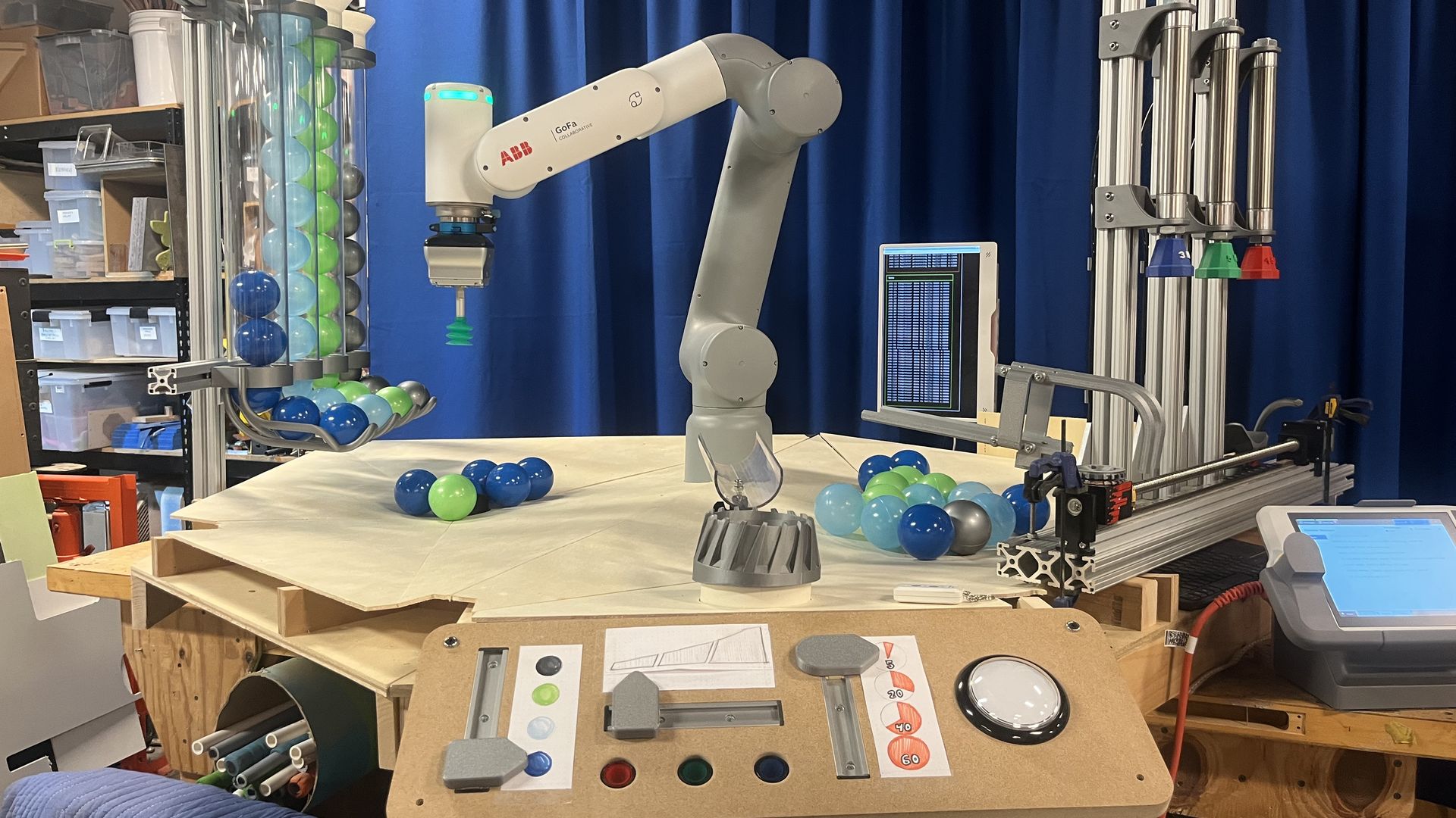 Industrial lab with a gray ABB GoFa collaborative robotic arm at a worktable. Blue curtain backdrop, monitor screen, and racks of multicolored plastic balls (blue, green) around the workspace.
