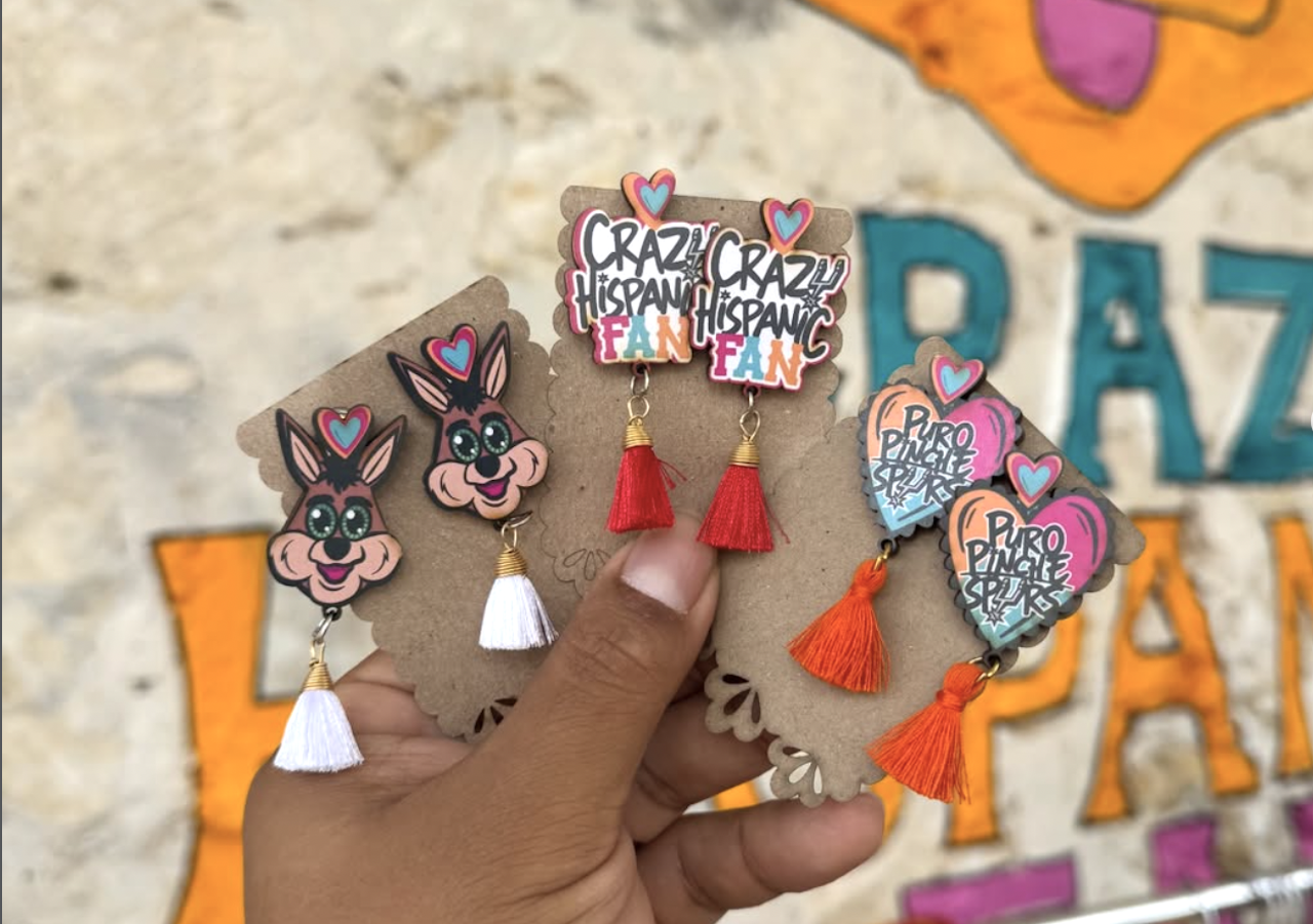 Hand holding a cardboard display of quirky earrings: bunny-face charms, heart accents, white, red, and orange tassels, and a "Crazy Hispanic Fan" badge, against a colorful mural backdrop.