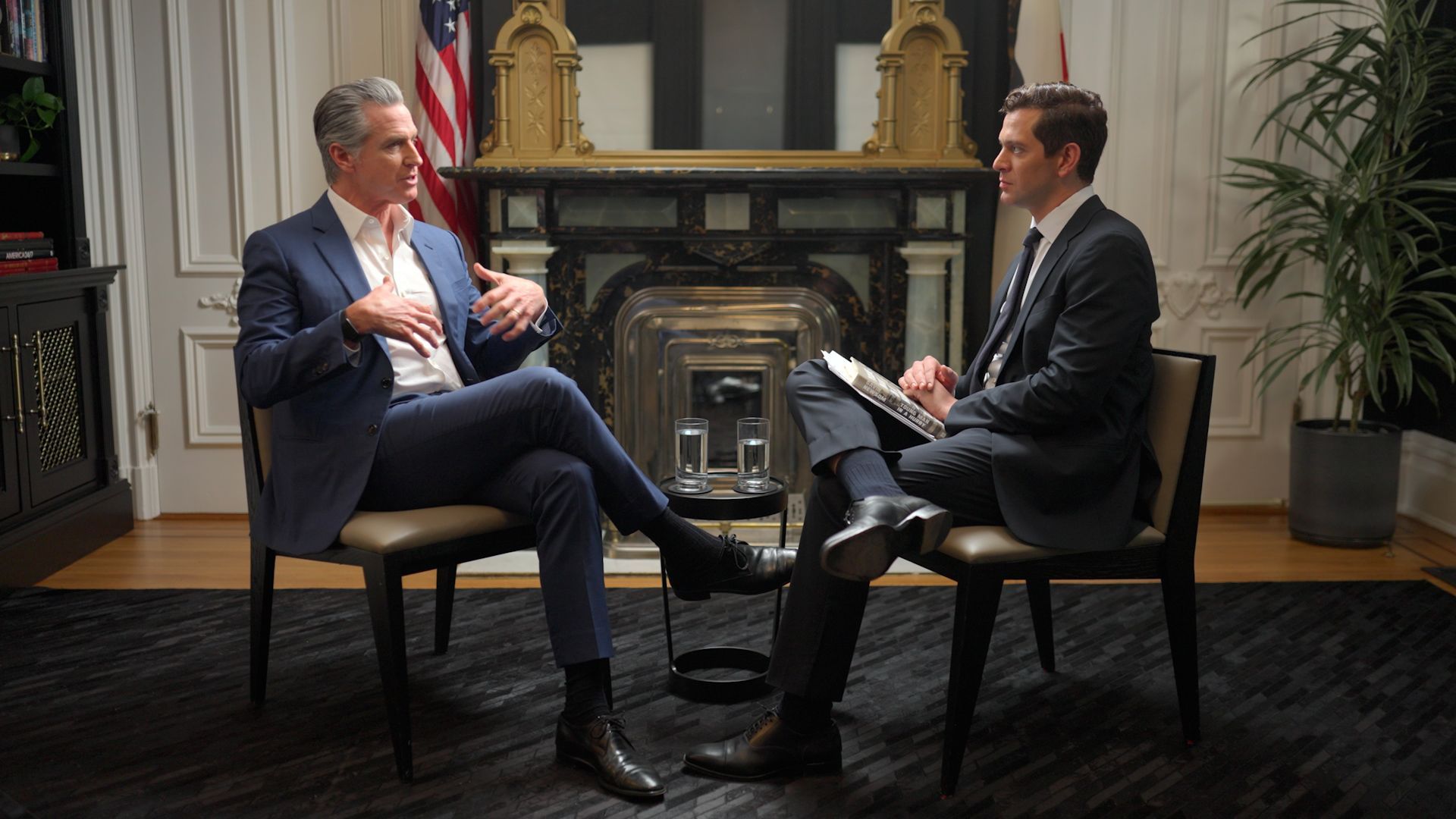 California Gov. Gavin Newsom and another man in a suit sit facing each other in a formal room, engaged in an interview, with a fireplace and U.S. flag in the background.