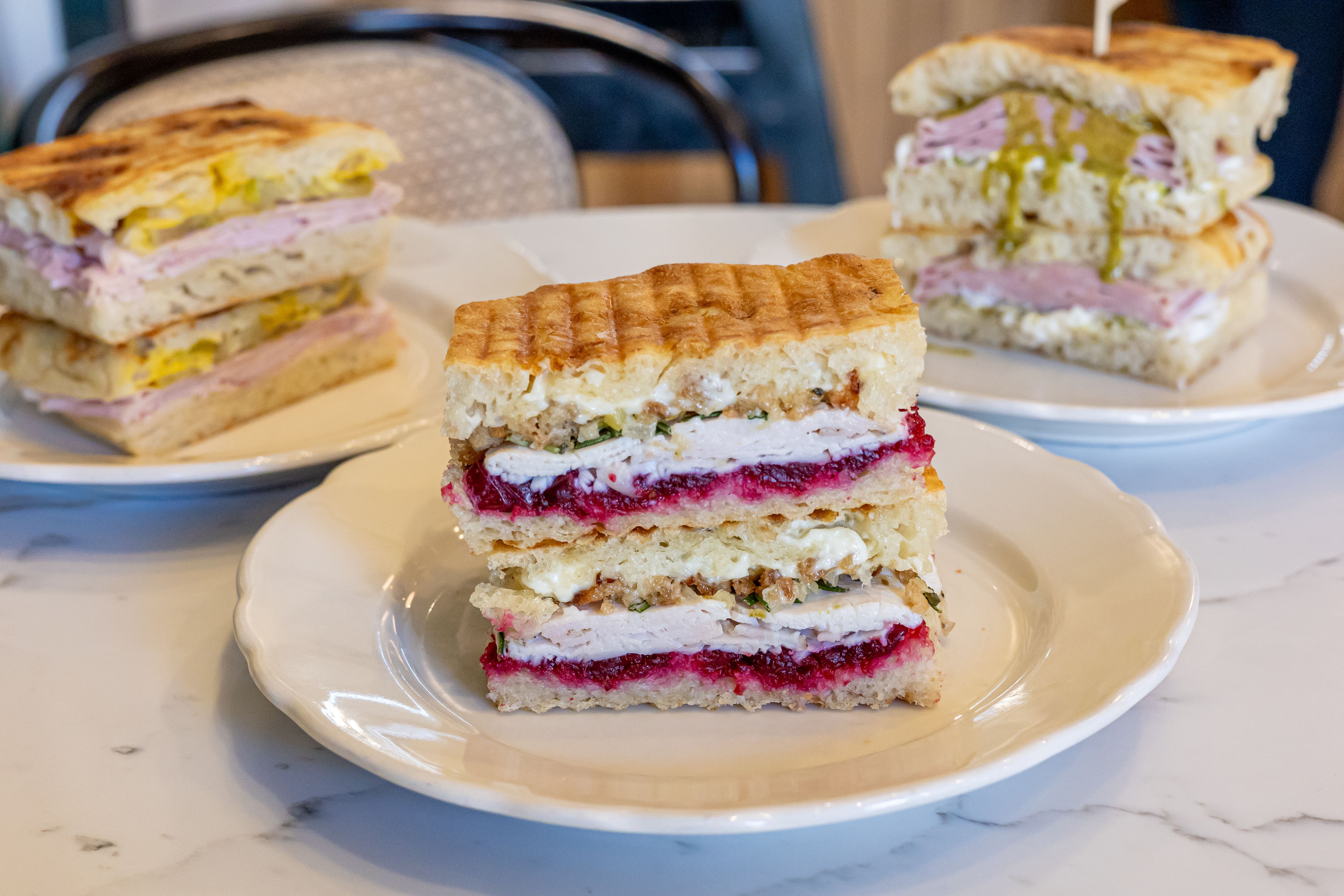 Three sandwiches on white plates on a marble table, each with focaccia bread and different fillings including turkey, cranberry sauce, ham, cheese, and mustard.