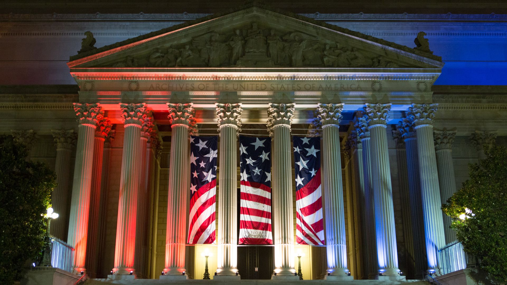 Night view of the U.S. National Archives building with tall Corinthian columns, red, white, and blue lighting, American banners between the columns, and a glowing entrance lamp.
