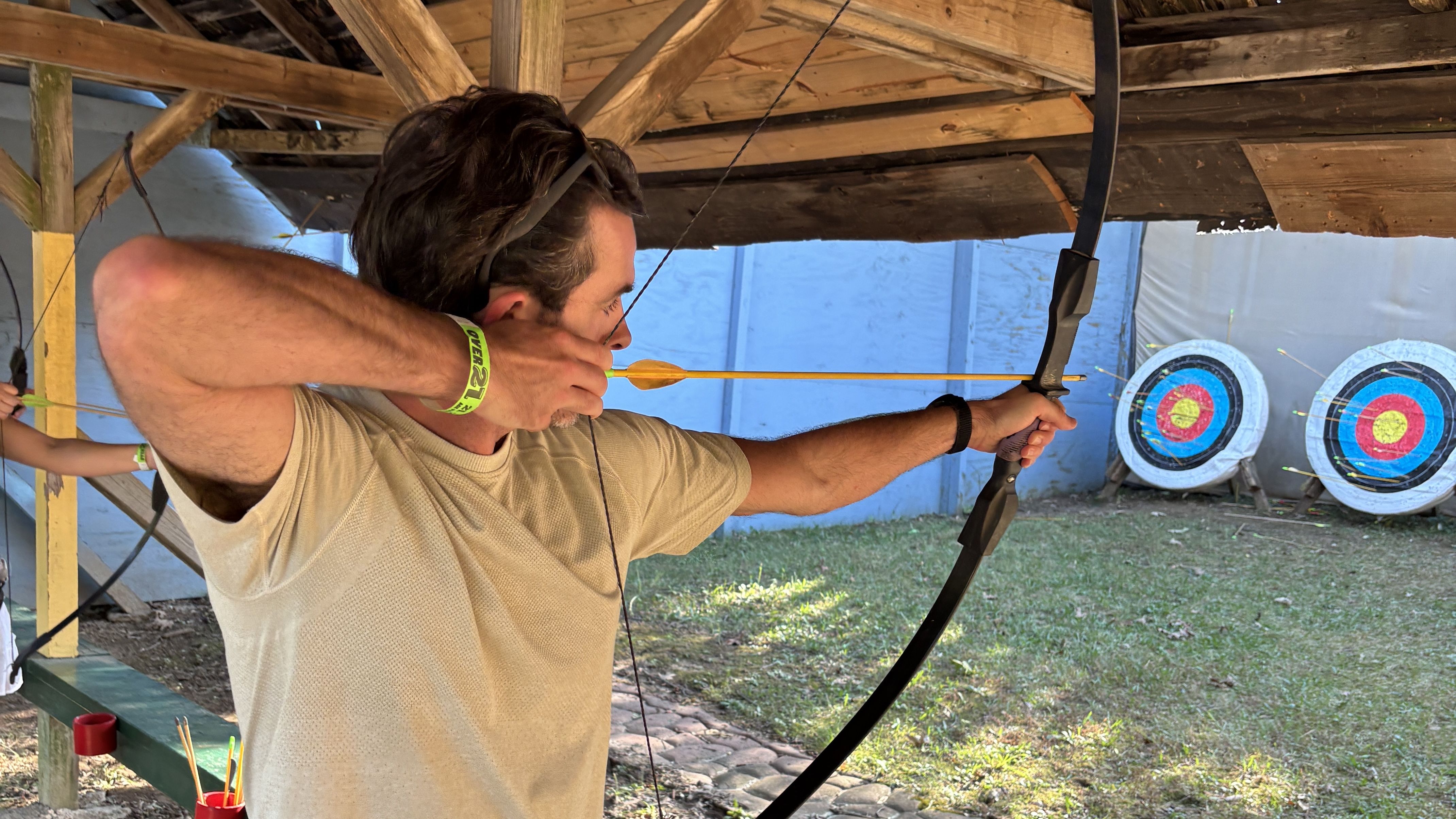 A man in a beige shirt draws a black bow at an outdoor archery range under a wooden shelter; three targets with blue, red, and yellow rings are visible in the background.