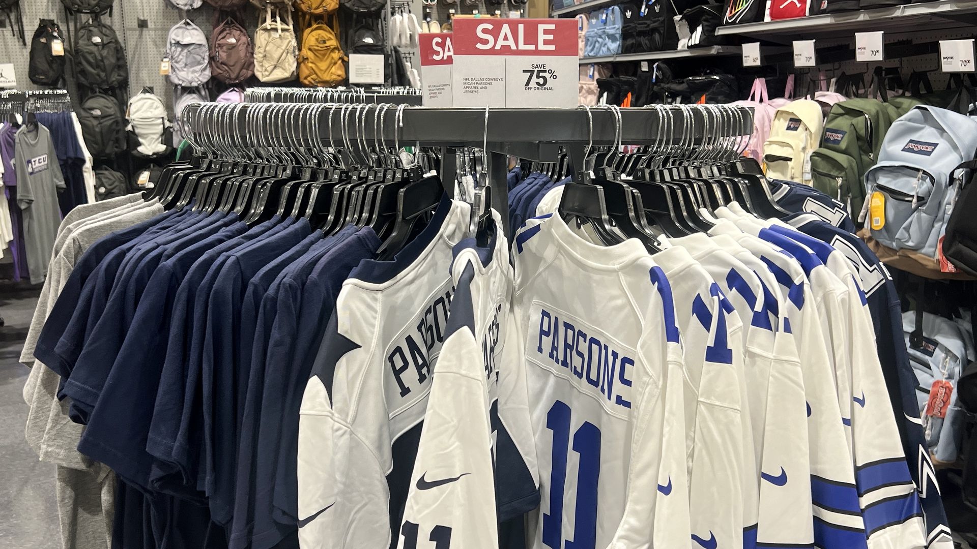 Rack of NFL Dallas Cowboys apparel including white and navy #11 Parsons jerseys and t-shirts in a store with a red sale sign advertising 75% off original prices.