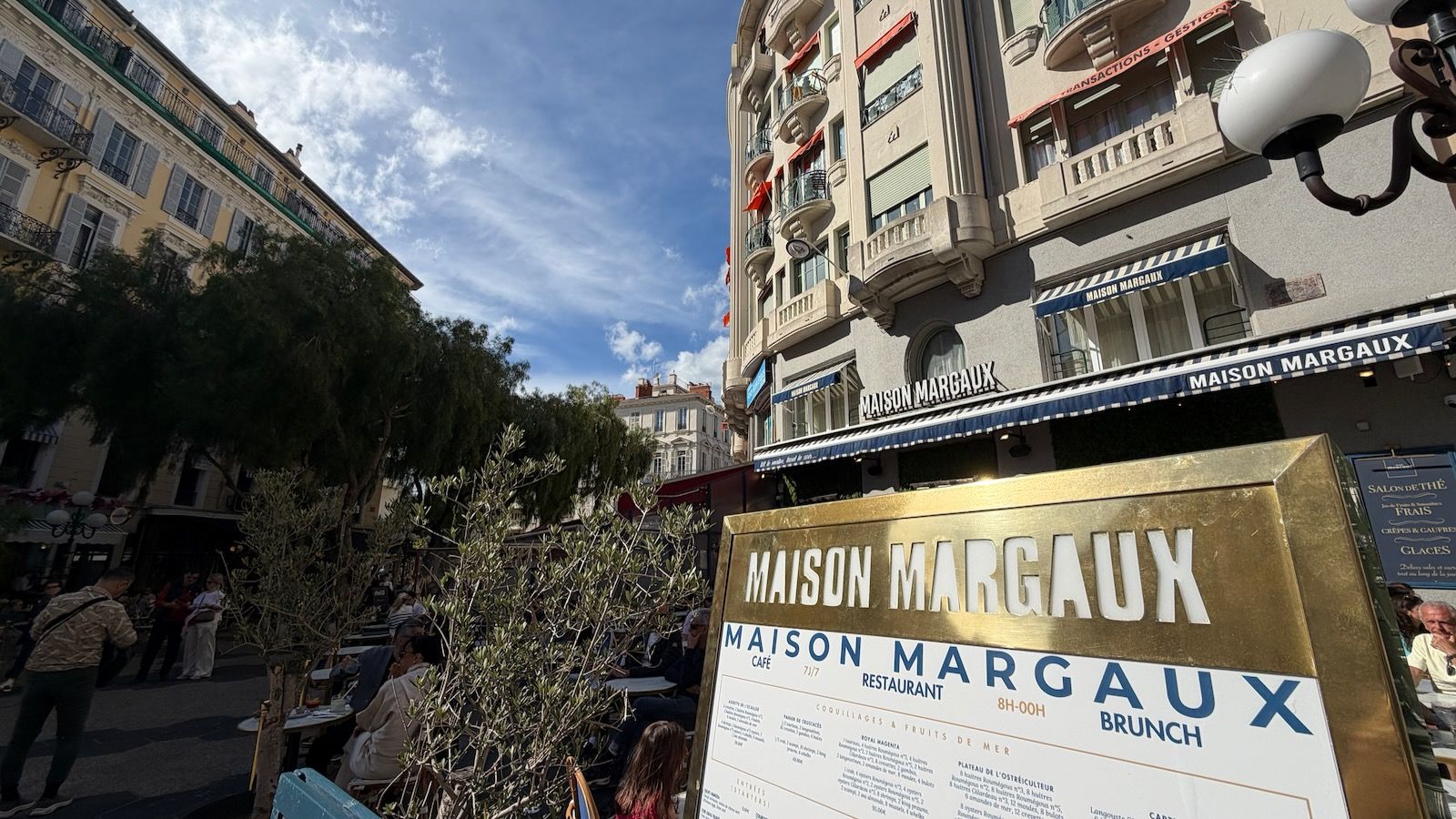 Sunny street scene with a large gold sign reading MAISON MARGAUX in the foreground; blue-and-white awnings over a curved building, outdoor café tables and pedestrians under a clear blue sky.