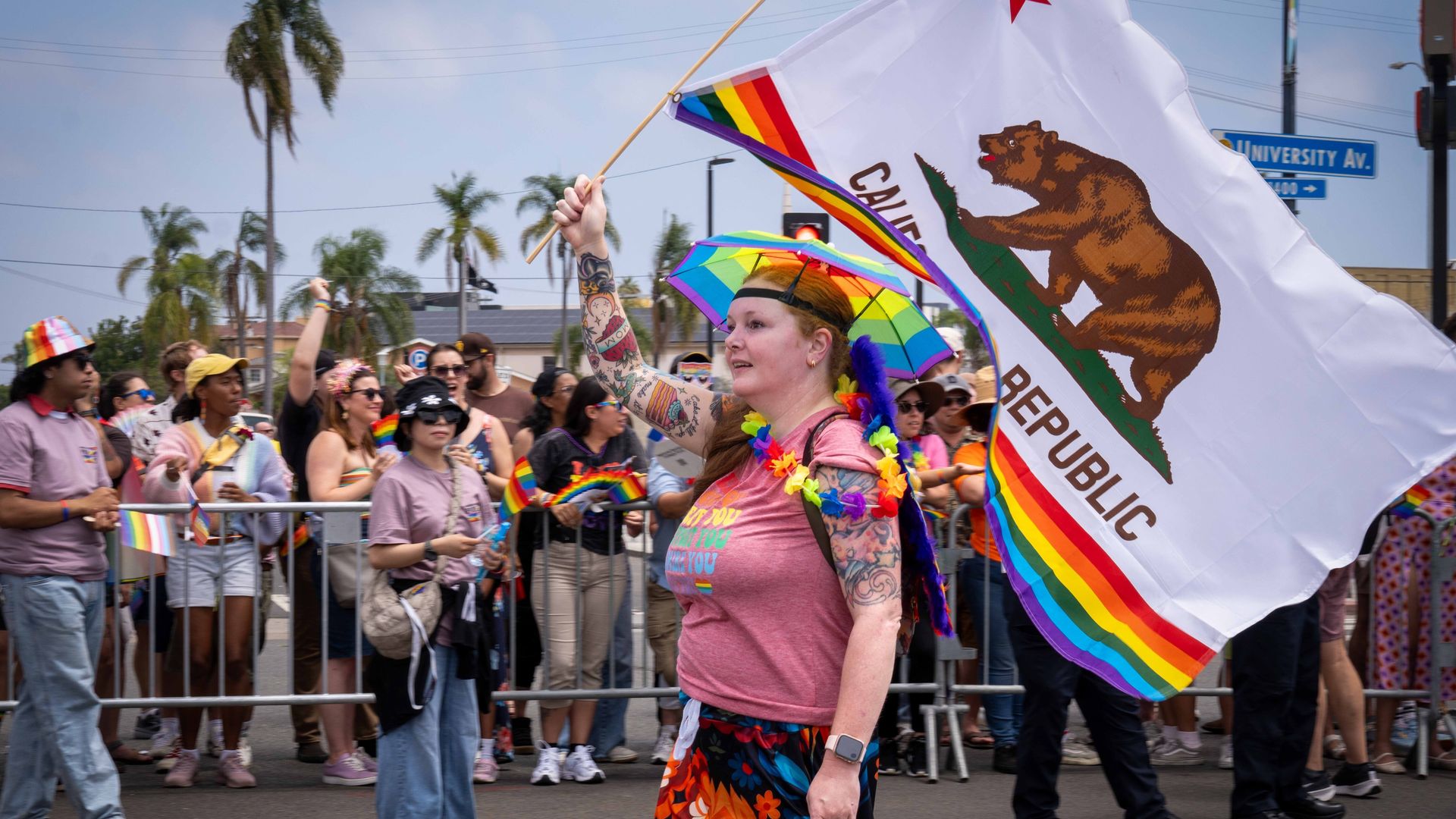 SAN DIEGO, CALIFORNIA, UNITED STATES - 2025/07/19: A marcher wearing a rainbow umbrella hat and colorful leis waves a California state flag modified with Pride colors. Spectators holding small rainbow flags line the street behind the barricades. Participants marched during the San Diego Pride Parade