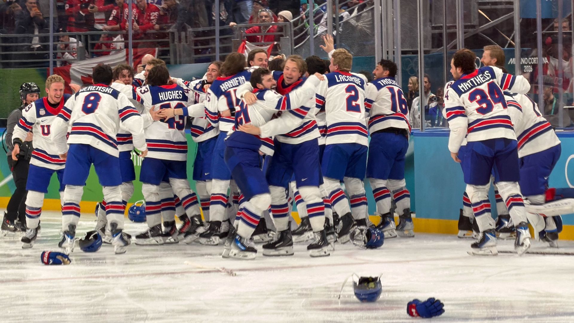 Ice hockey team in white, blue, and red uniforms celebrating on ice rink, hugging and cheering, with crowd waving American flags in background.