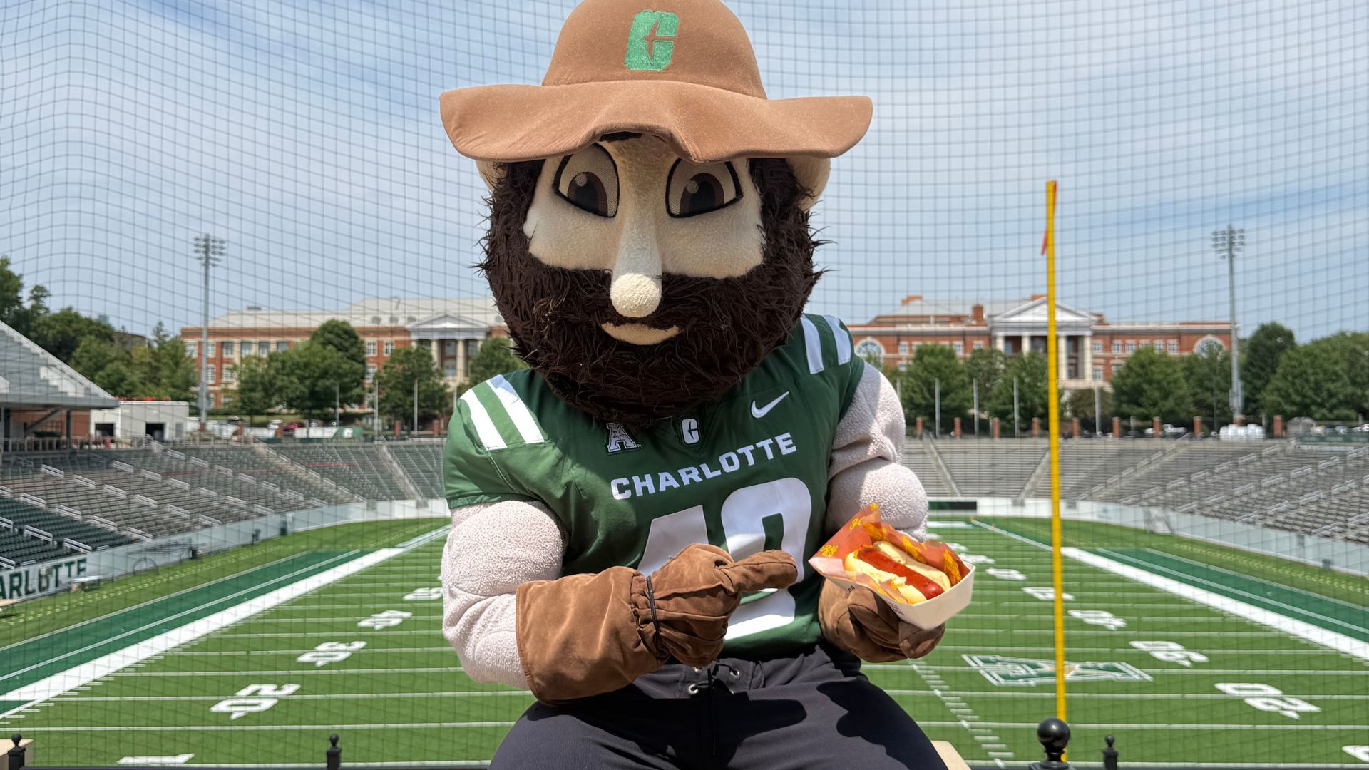 Mascot with a tan hat and green C, bearded face, wearing Charlotte jersey, holding hot dogs on a football field; empty stands and campus buildings in the background.